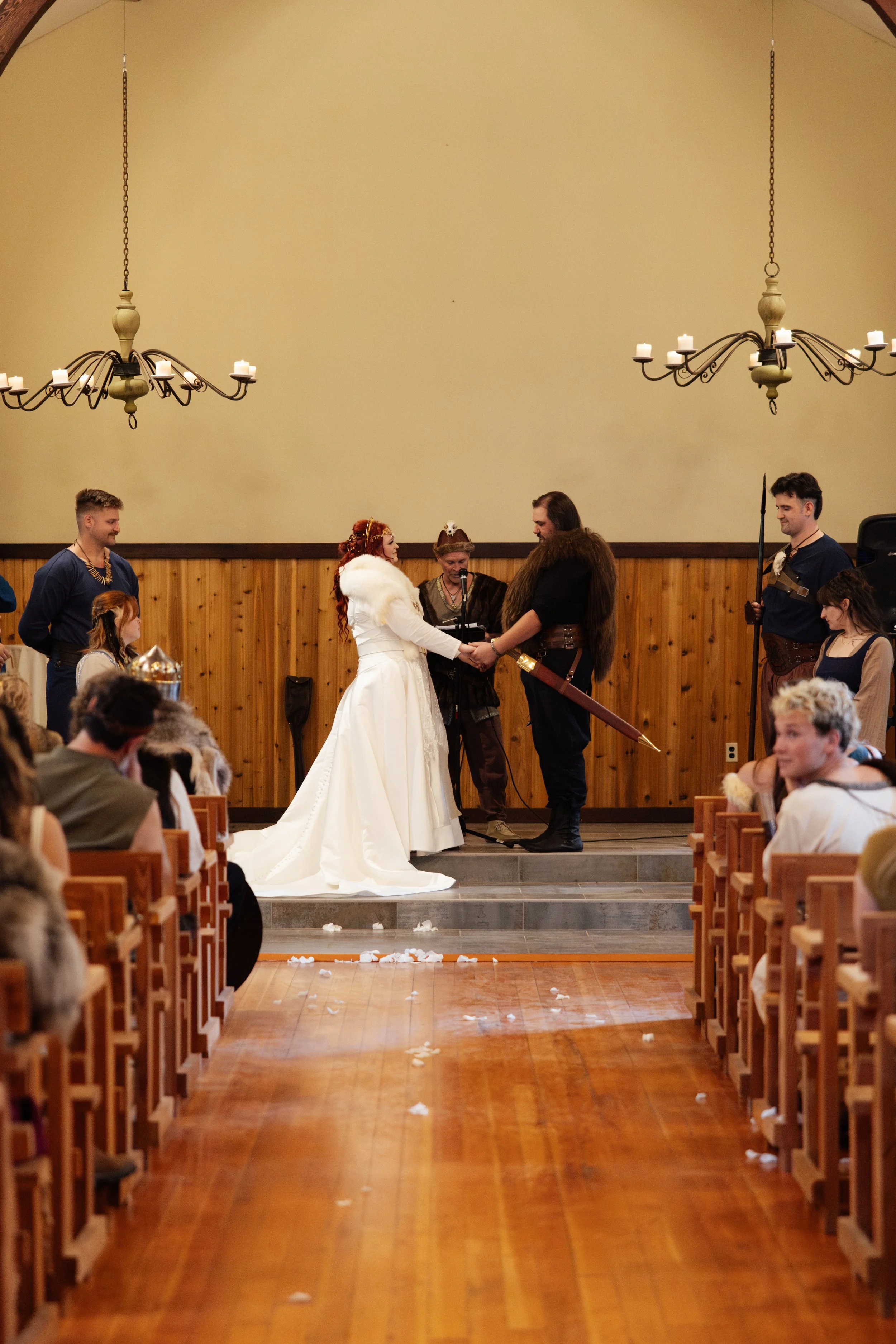 A wedding ceremony in a church with a bride and groom holding hands in front of an officiant. The bride is wearing a white gown and the groom is dressed in medieval or fantasy-themed attire with a fur cloak and sword. Guests are seated on wooden pews