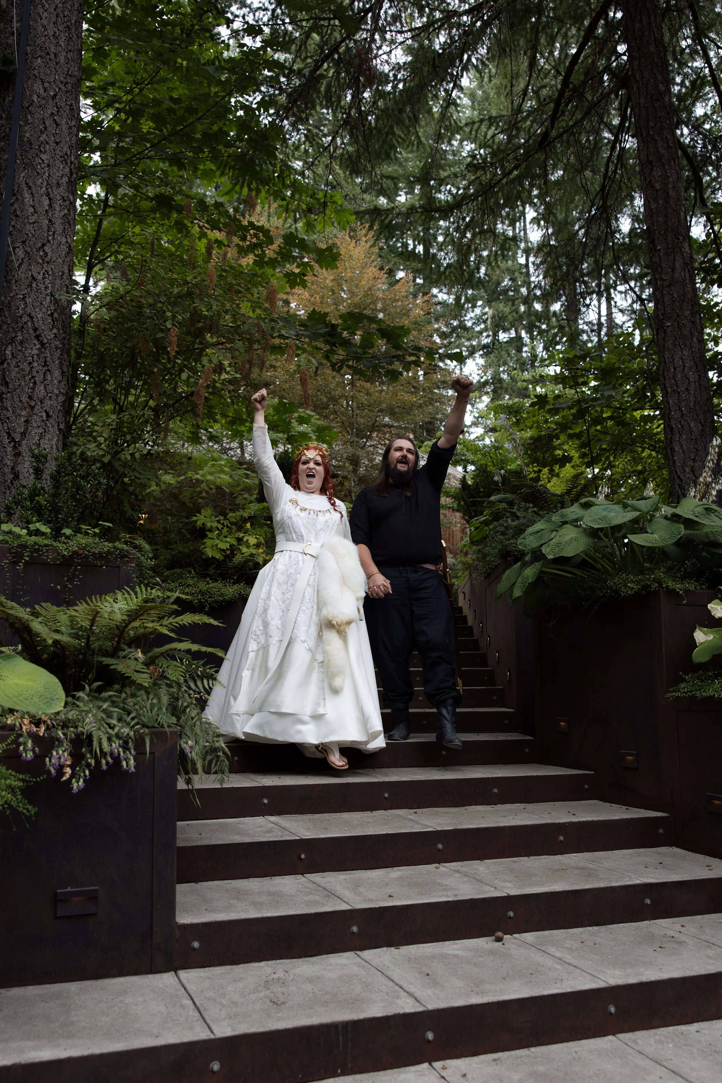 A joyful couple holding hands and celebrating on outdoor steps amid lush greenery and tall trees. The woman wears a white wedding dress and the man a black shirt and black pants.