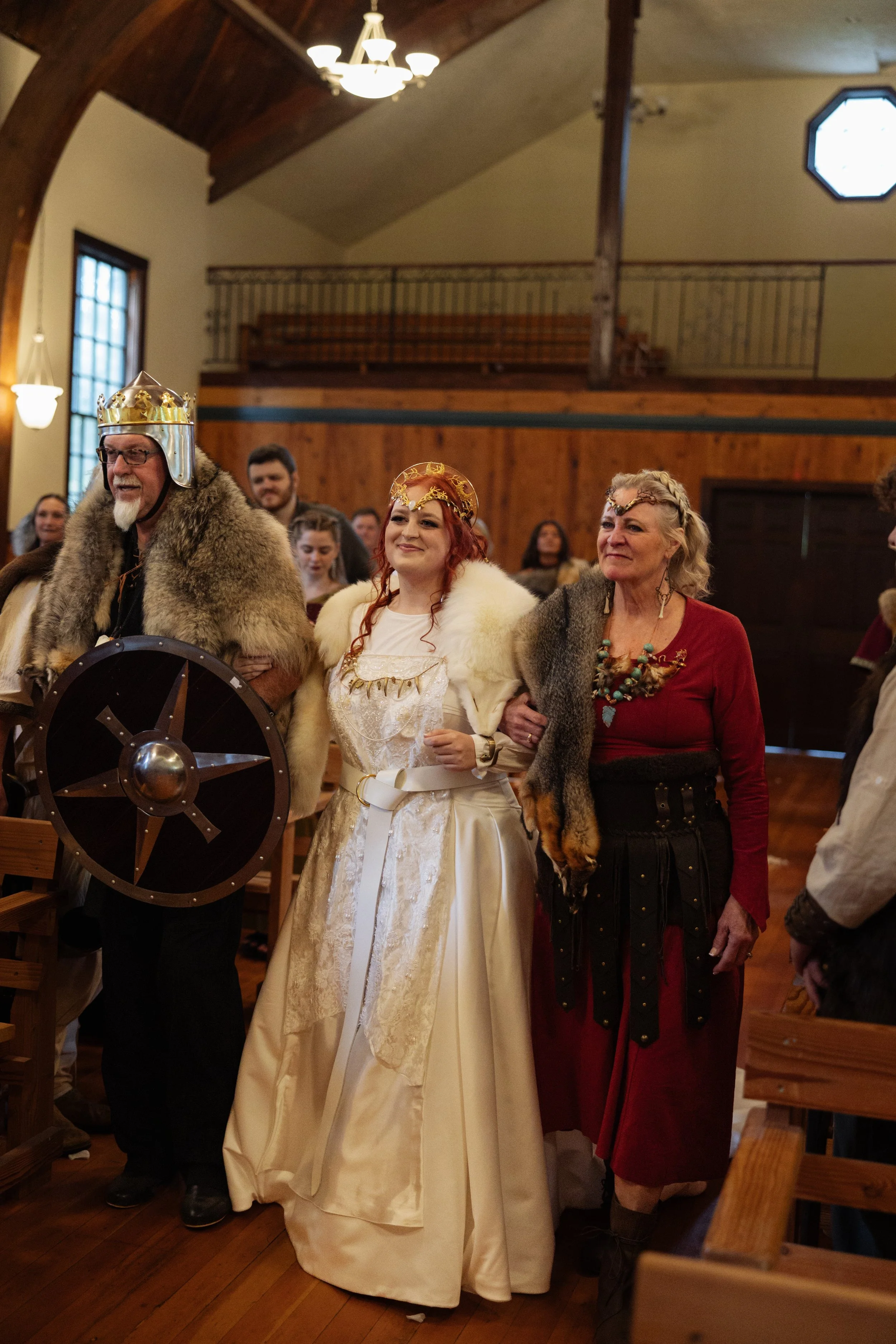 People dressed in medieval costumes, including a man with a crown and fur cloak, and women with crowns and fur shawls, in a wooden hall with benches and warm lighting.
