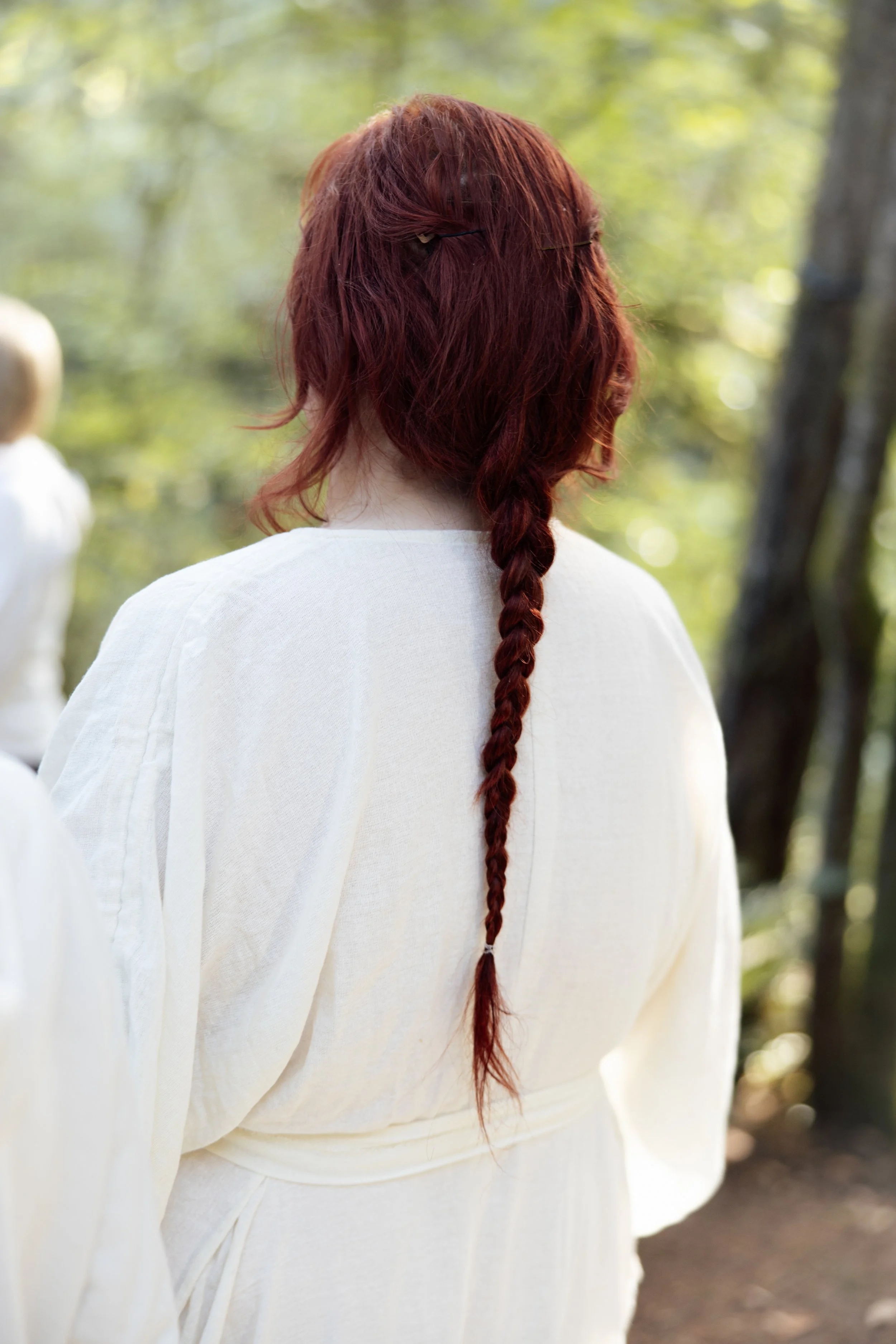 Back of a woman with long, wavy red hair in a braid, standing outdoors wearing a white top, with trees and sunlight in the background.