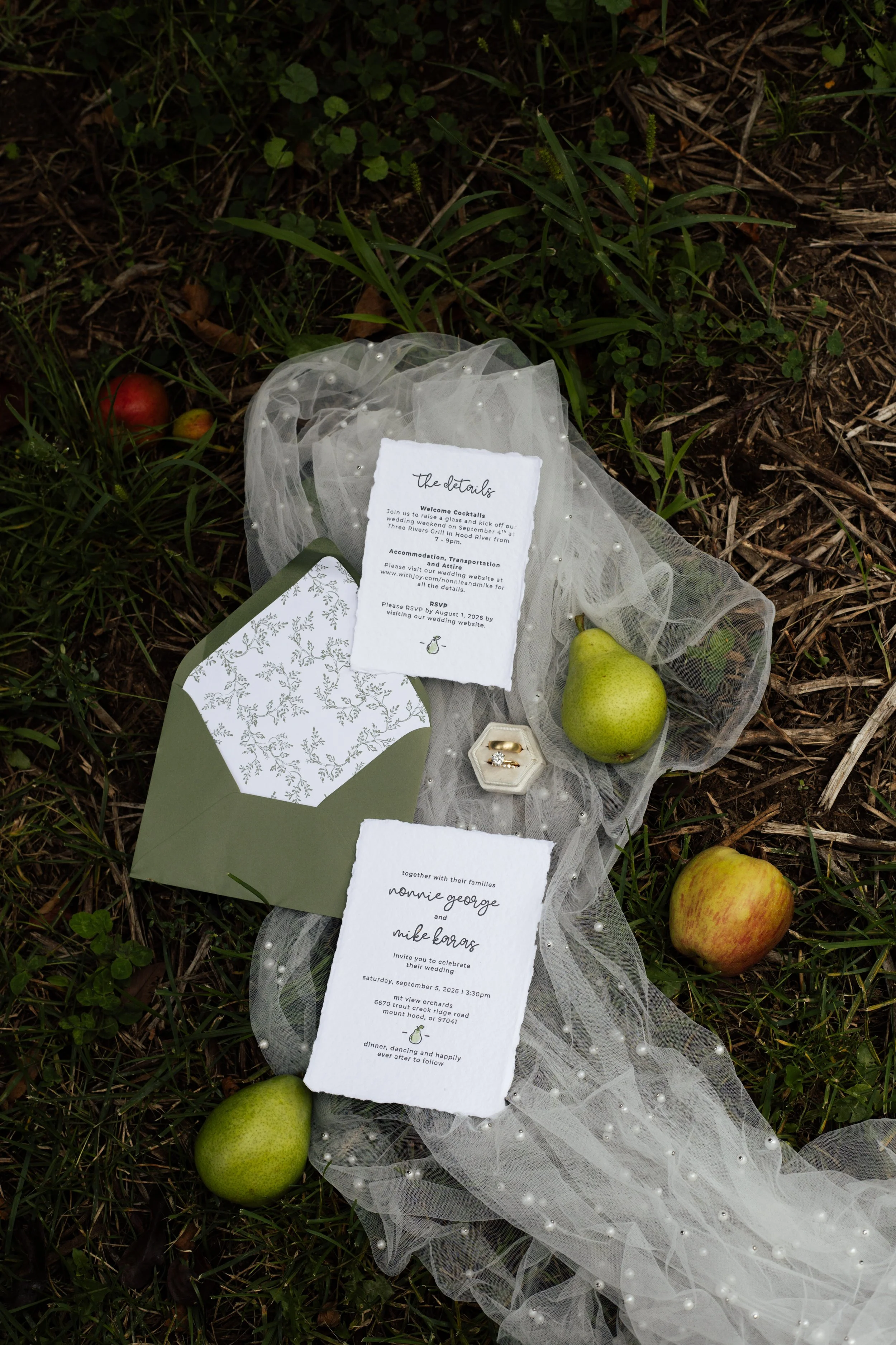 Wedding invitation cards with a white tulle fabric decorated with pearls, green apples, and scattered apples on grass.