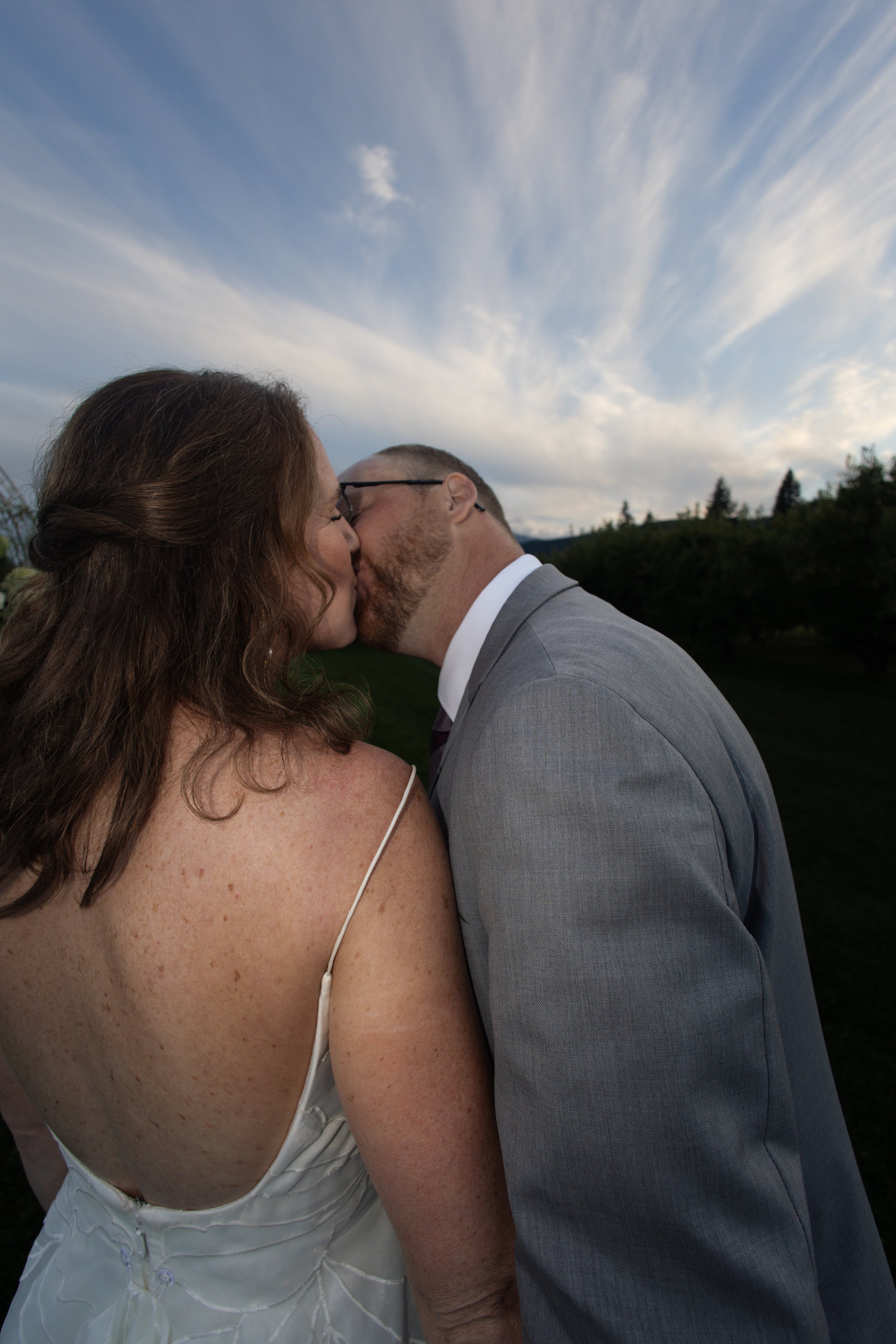 A close-up of a couple sharing a kiss outdoors, with a cloudy blue sky and trees in the background.