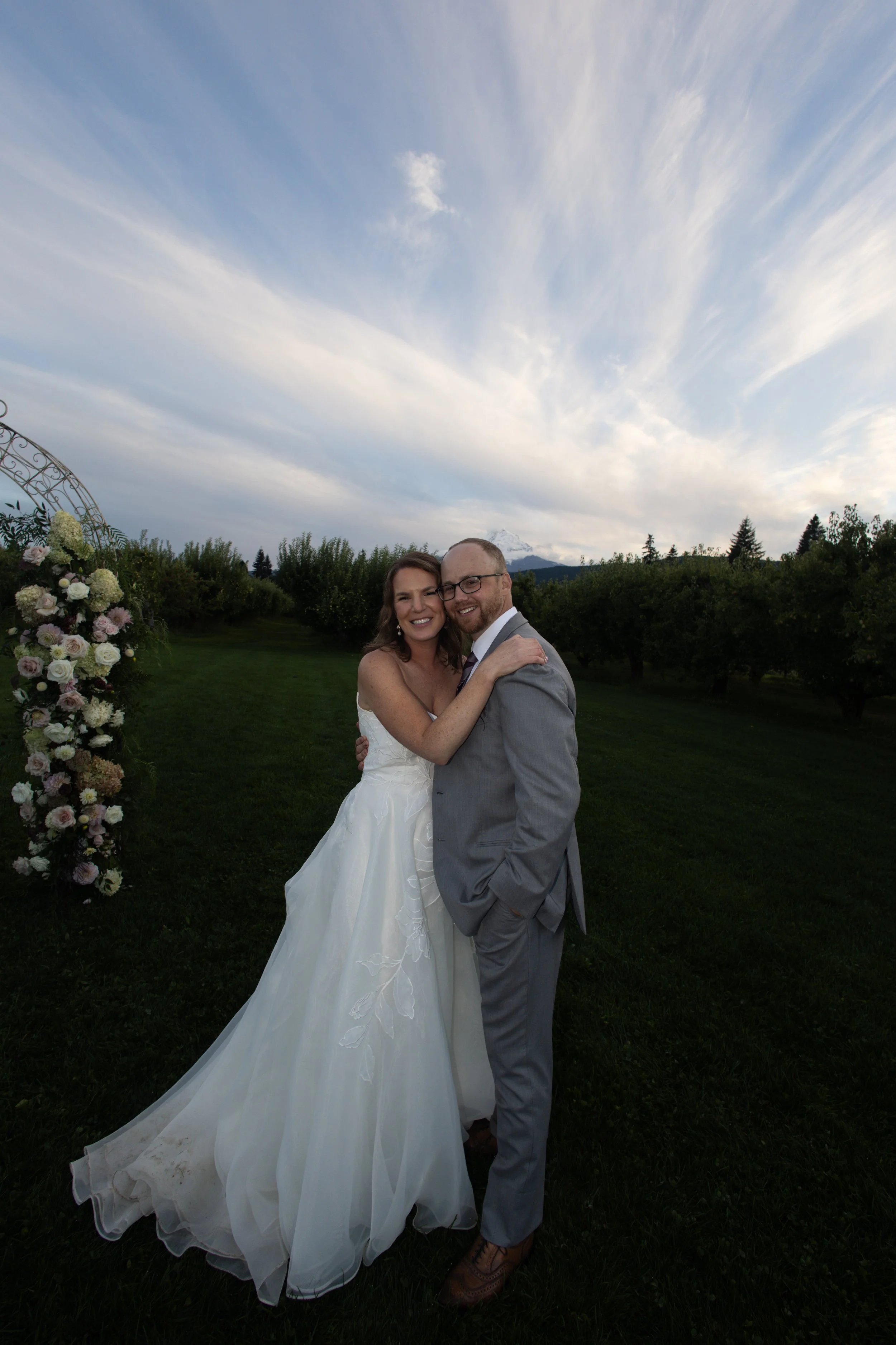 A happy couple in wedding attire embracing outdoors on a grassy field with a cloudy sky and mountains in the background. The woman is wearing a white wedding gown, and the man is in a gray suit. There is a floral decoration arch on the left side of t