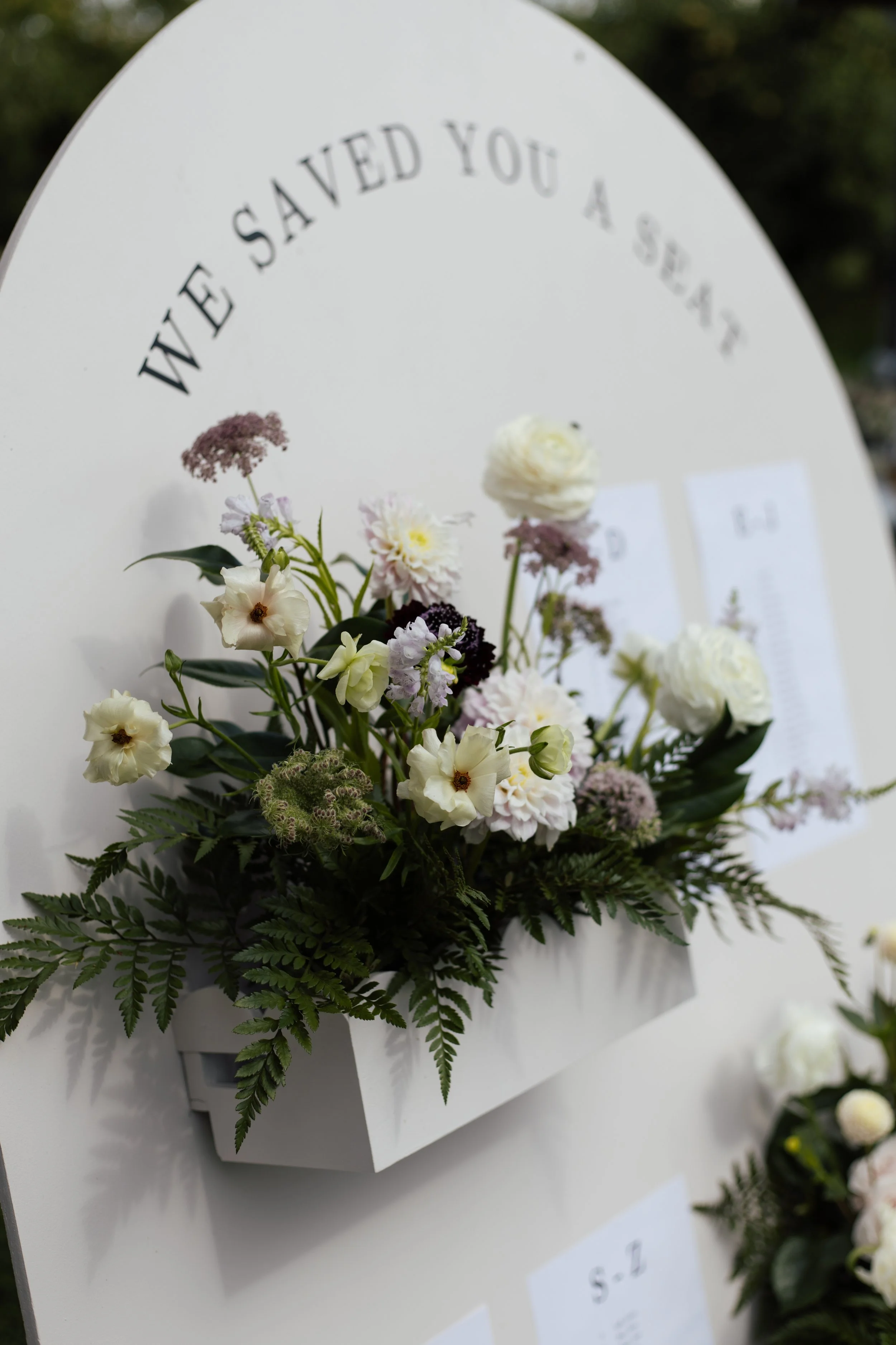 A floral arrangement with white and purple flowers and greenery in front of a white card with the words "We have saved you a seat" written on it.