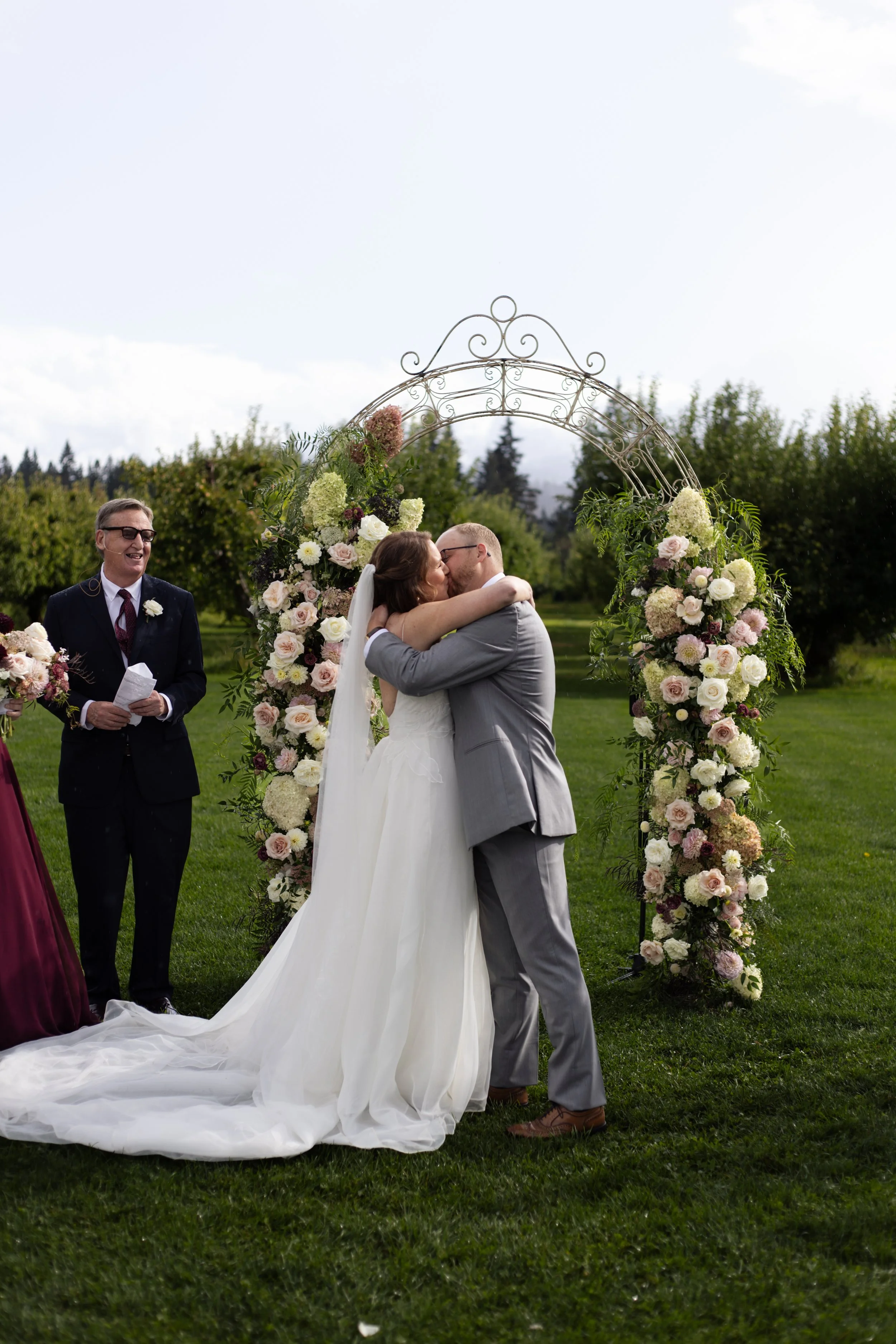 A bride and groom kissing at their outdoor wedding ceremony, framed by a floral arch with pink and white flowers. The officiant stands nearby, smiling and holding a paper, as green trees and cloudy sky are in the background.