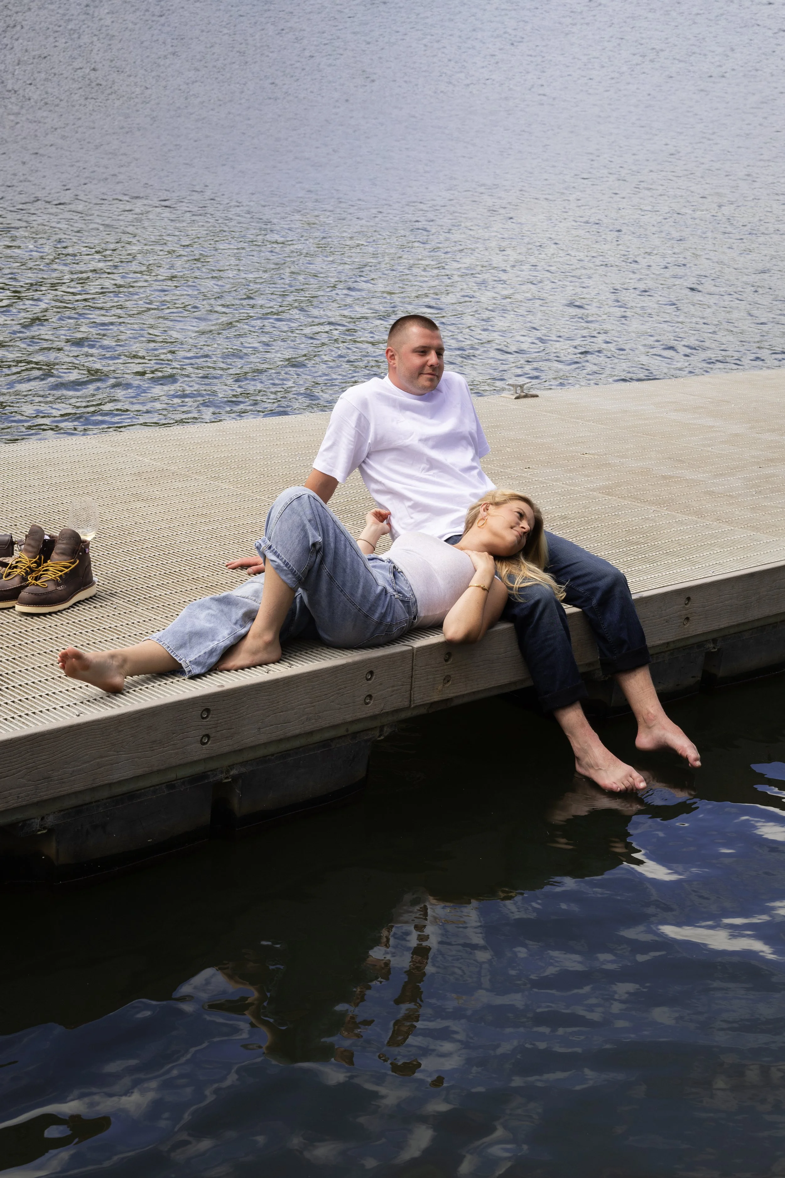 A man and a woman relaxing on a wooden dock over water, with the woman lying down and the man sitting behind her, both barefoot, with a pair of boots and an empty glass nearby.