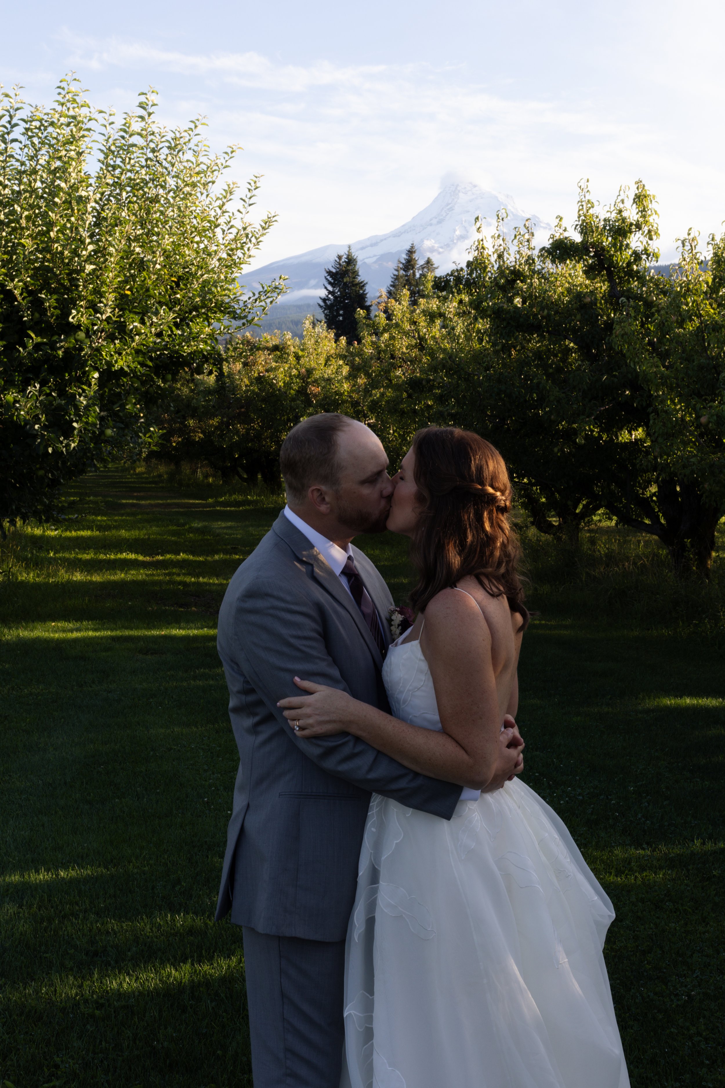 A couple dressed in wedding attire kissing outdoors in a grassy area with trees. A mountain with a snow-covered peak is in the background, under a partly cloudy sky.