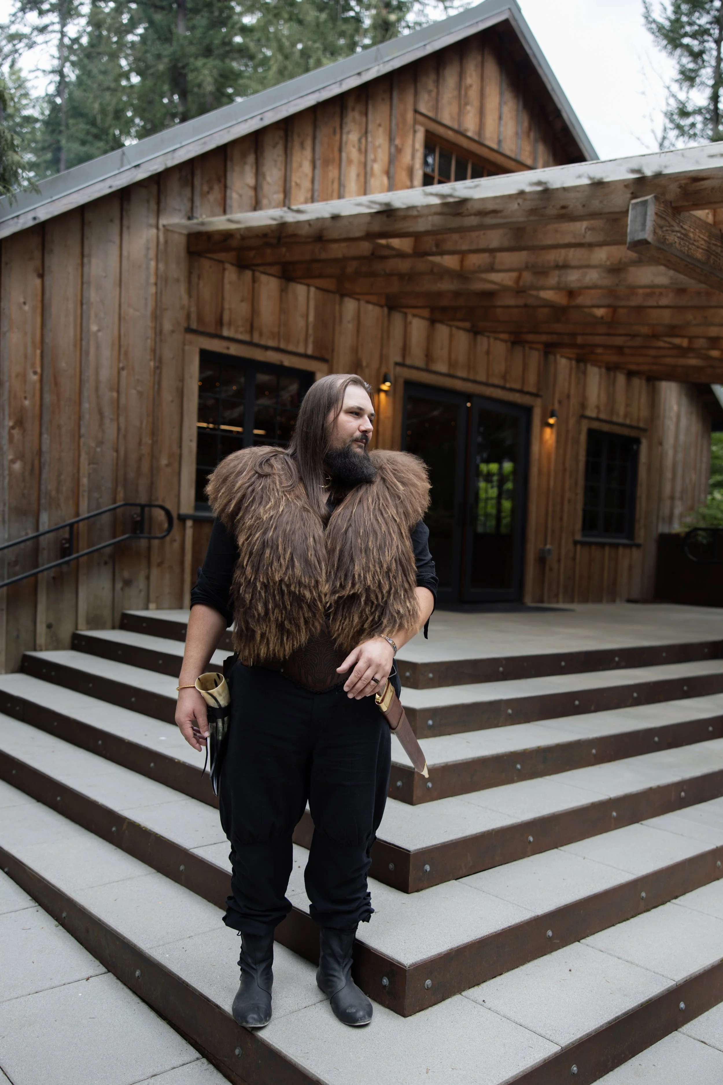 A person standing on stairs in front of a wooden building with a metal roof, dressed in black pants, boots, and a brown fur vest.