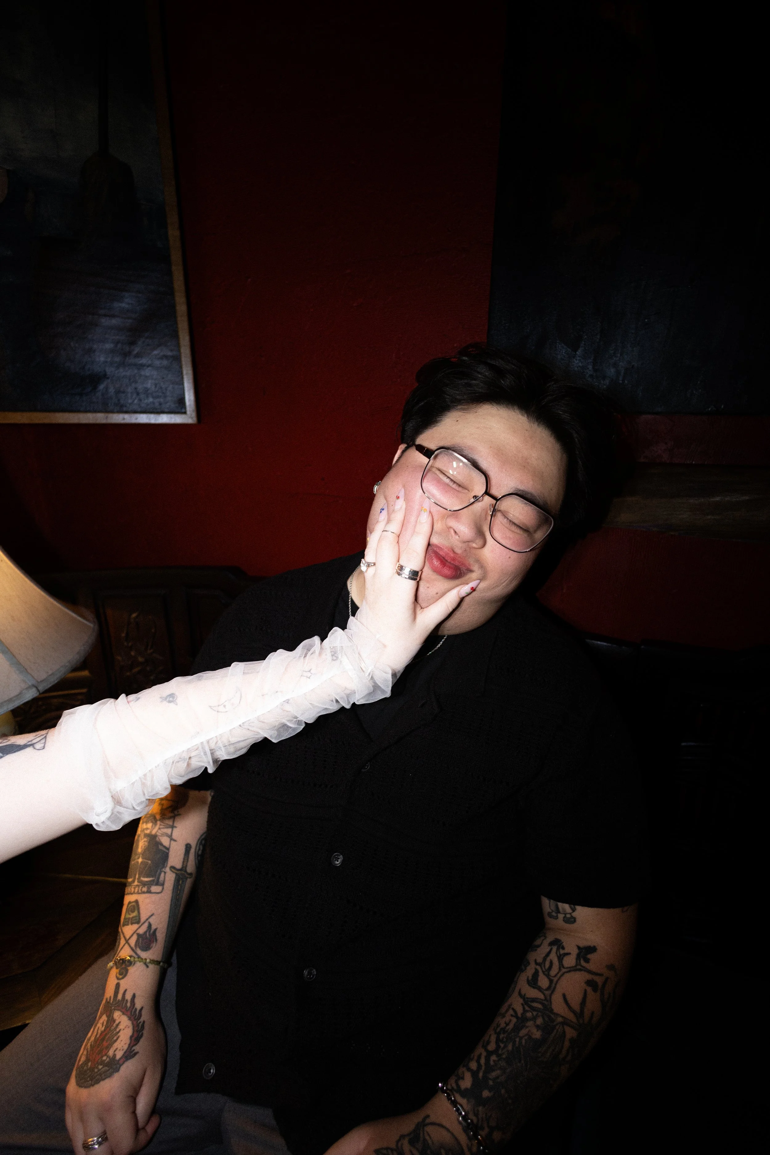 A woman with glasses and tattoos on her arms is smiling with her hand on her cheek. To her left, a person with light skin, light-colored nails with colorful designs, and some rings is touching her face. The background includes dark walls with artwork