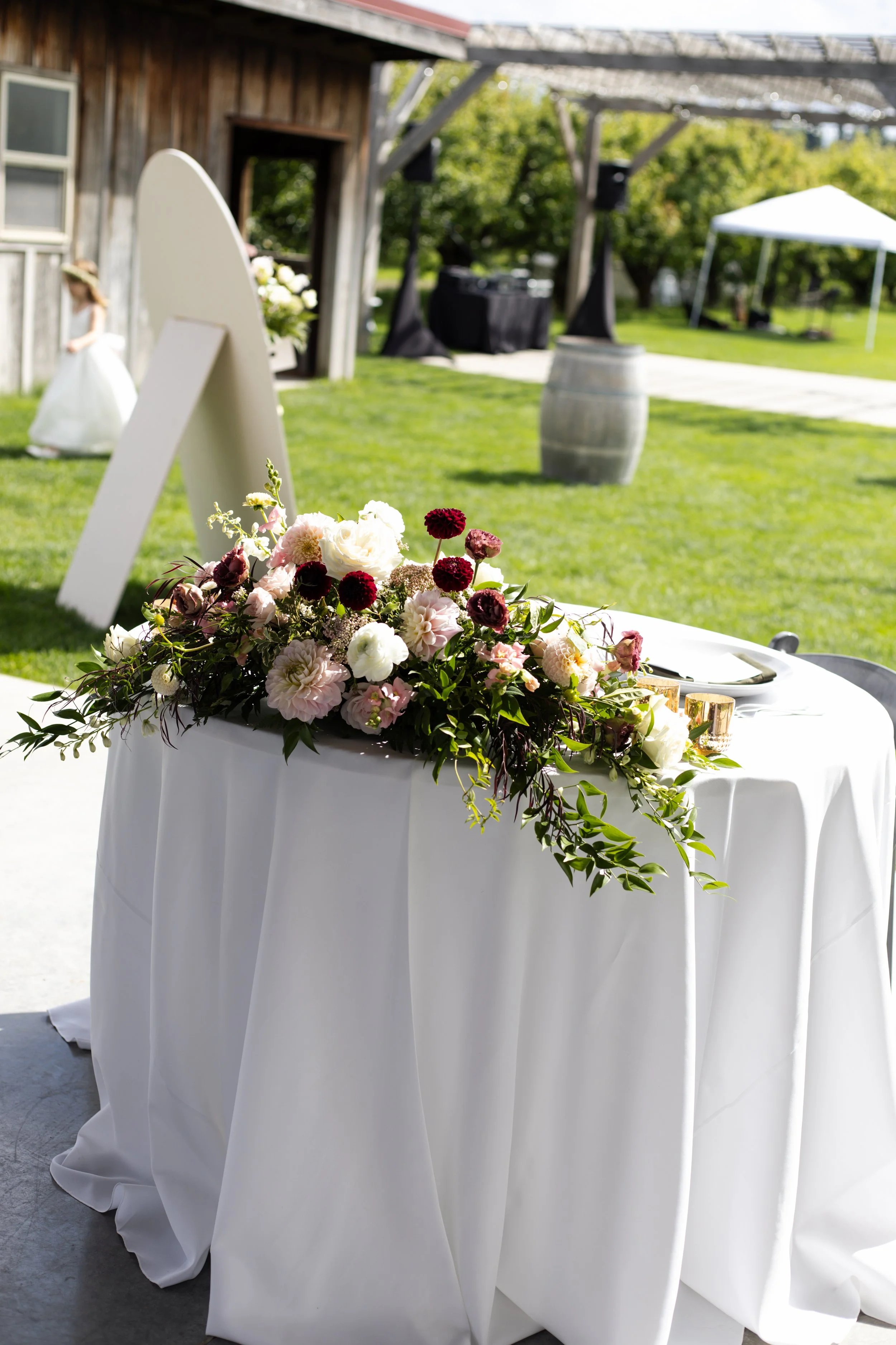 Wedding reception setup with a cake table and floral centerpiece, outdoor setting, with a barn and DJ equipment in the background.