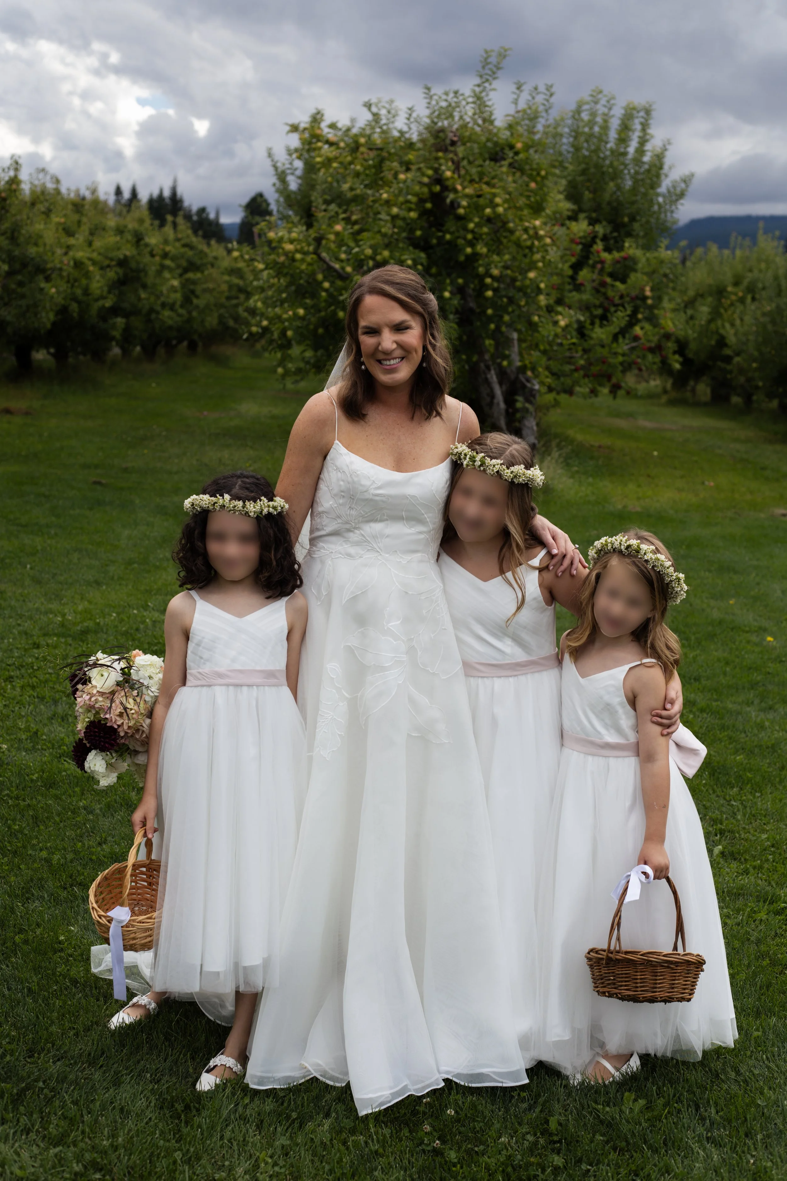 A woman in a white dress smiling with four young girls in white dresses with floral headbands, standing on a grassy field with trees and cloudy sky in the background.