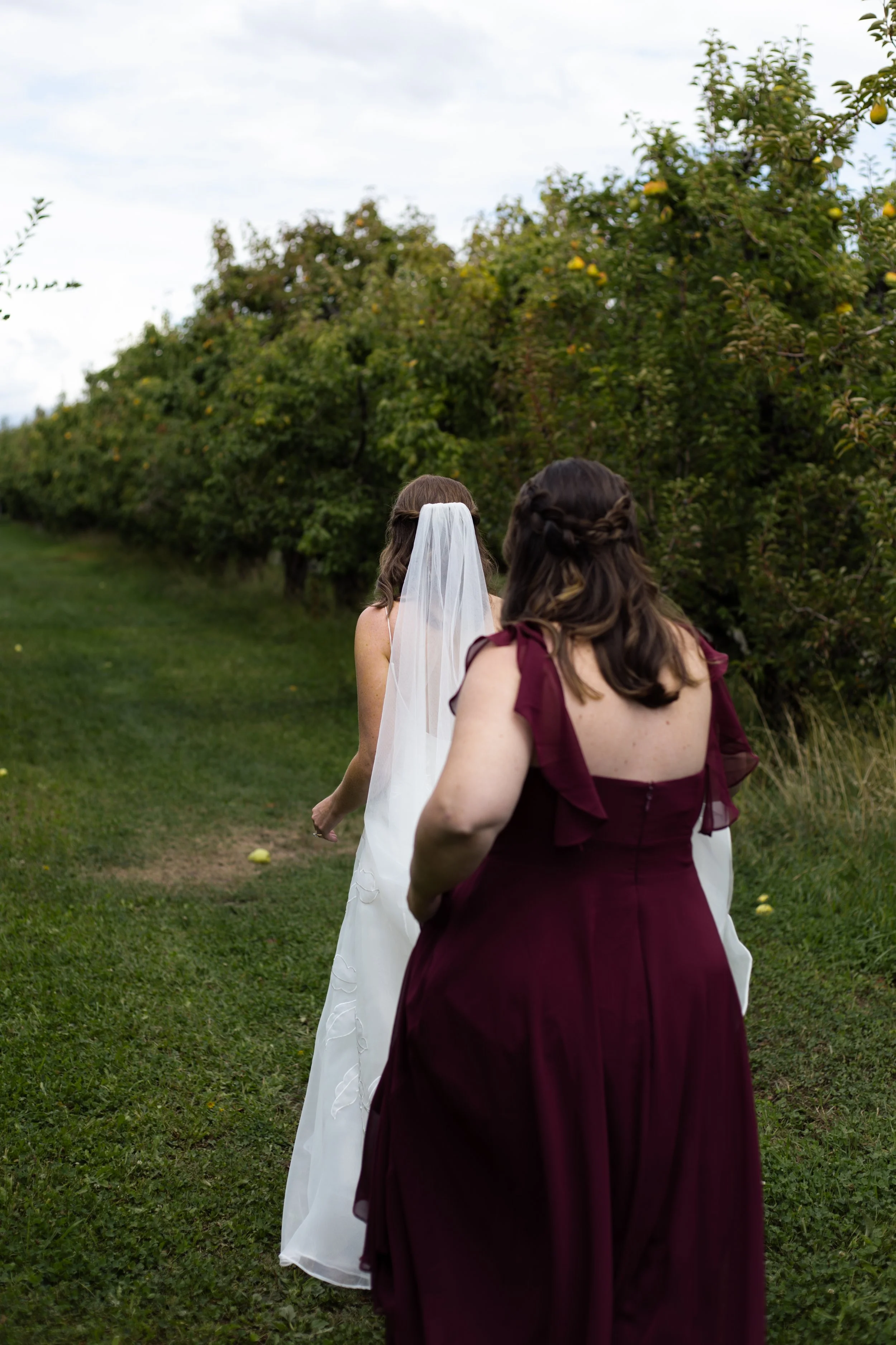 Two women walking outdoors in a grassy area with trees. One woman is dressed as a bride in a white wedding gown and veil, while the other woman is wearing a burgundy dress.