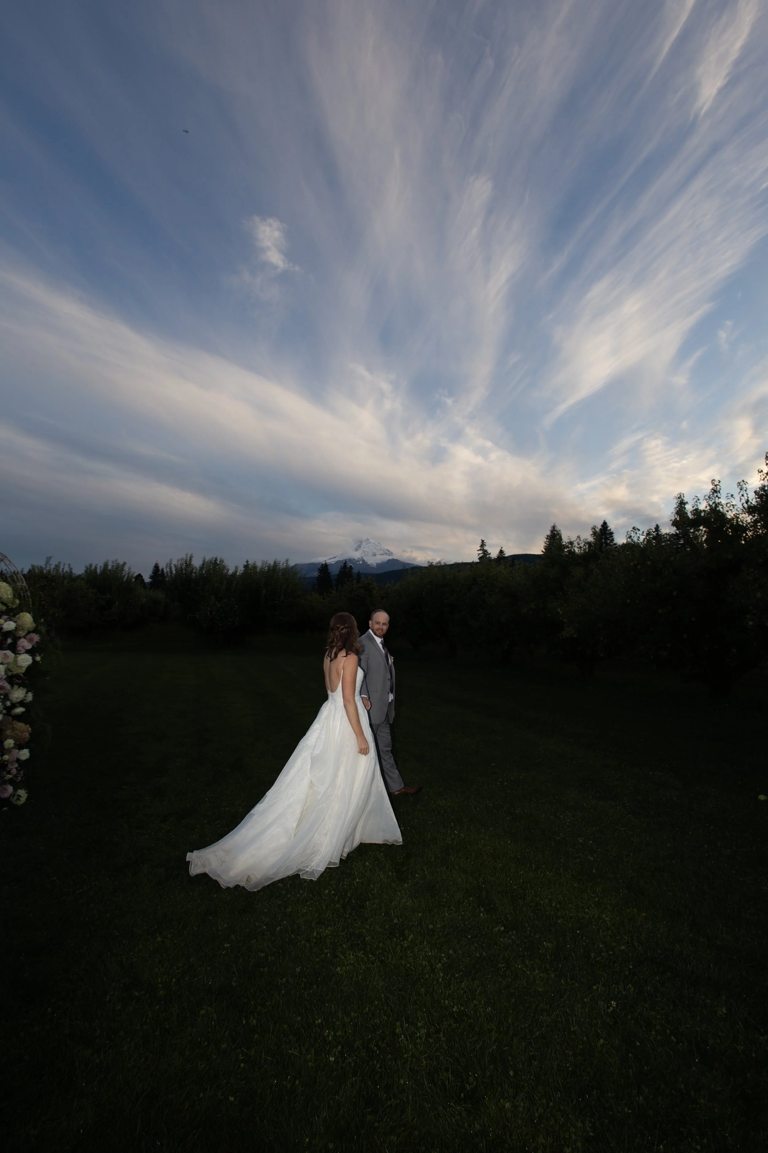 A bride and groom walking outdoors at sunset, with a mountain and trees in the background.