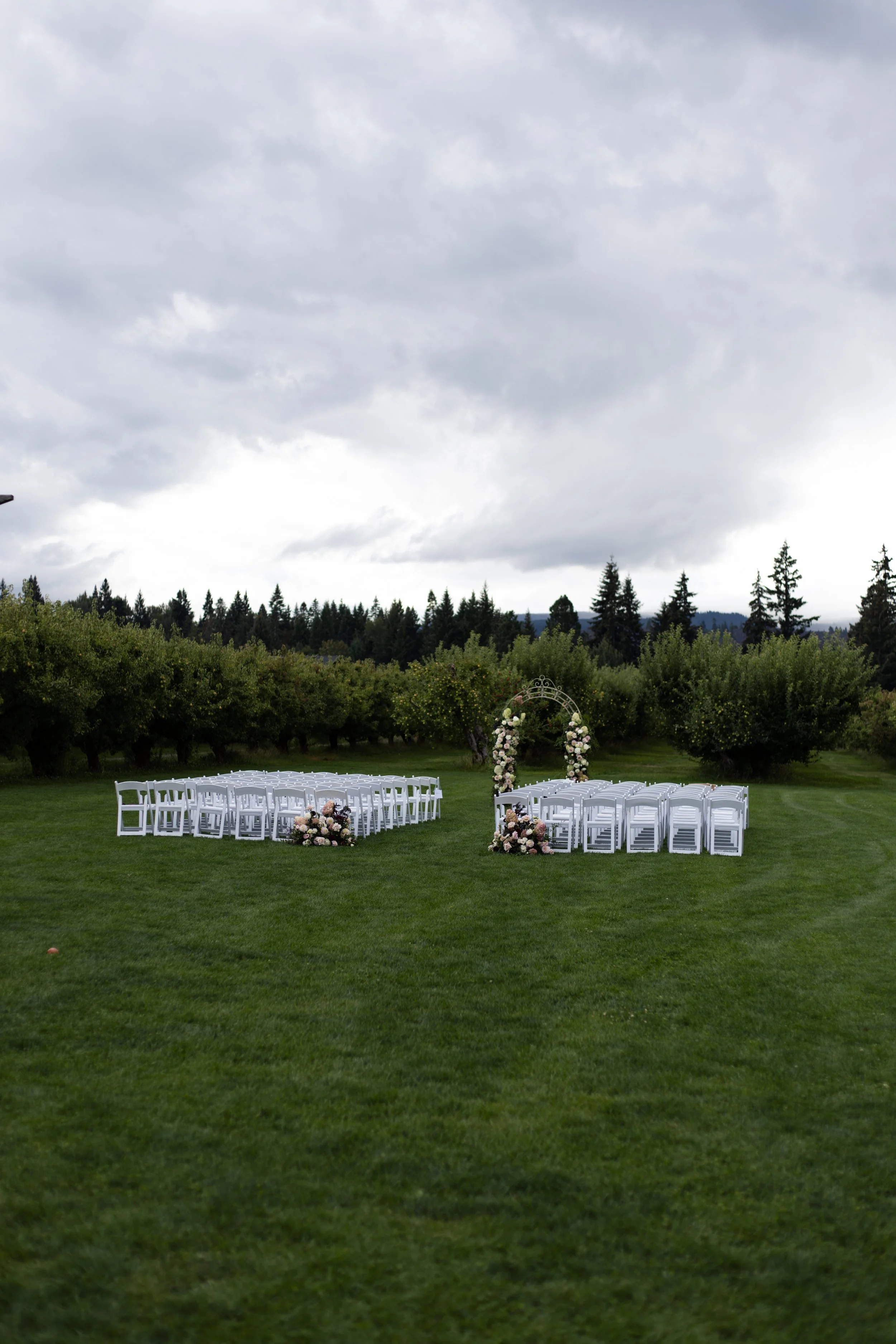 Outdoor wedding setup with white chairs arranged in two sections facing an arched floral gateway on a grassy lawn, surrounded by trees and under a cloudy sky.