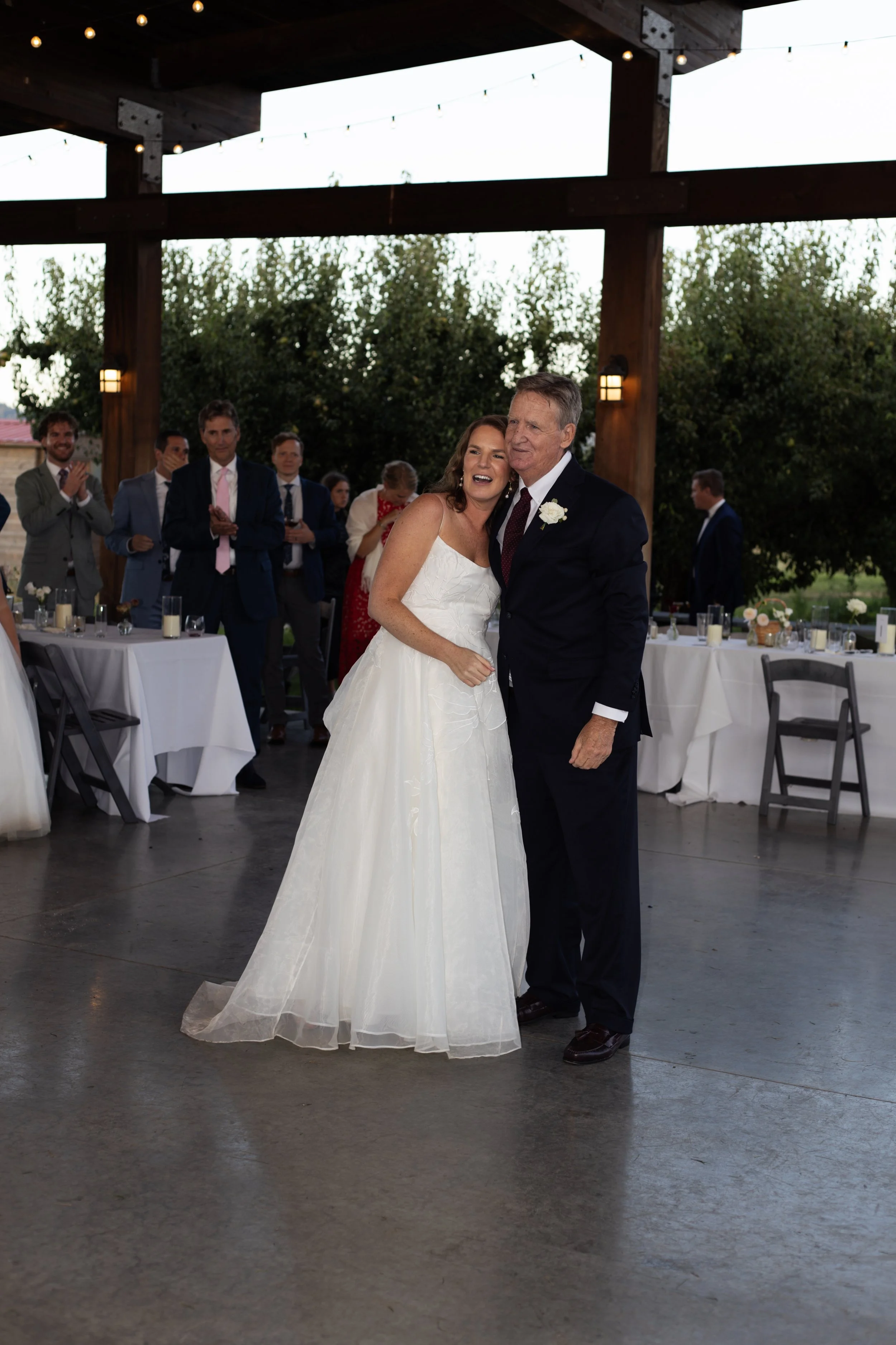 A bride and an older man, likely her father, hugging during a wedding reception. The bride is wearing a white wedding gown, and the man is dressed in a dark suit with a boutonnière. Several guests are clapping and smiling in the background under a wo