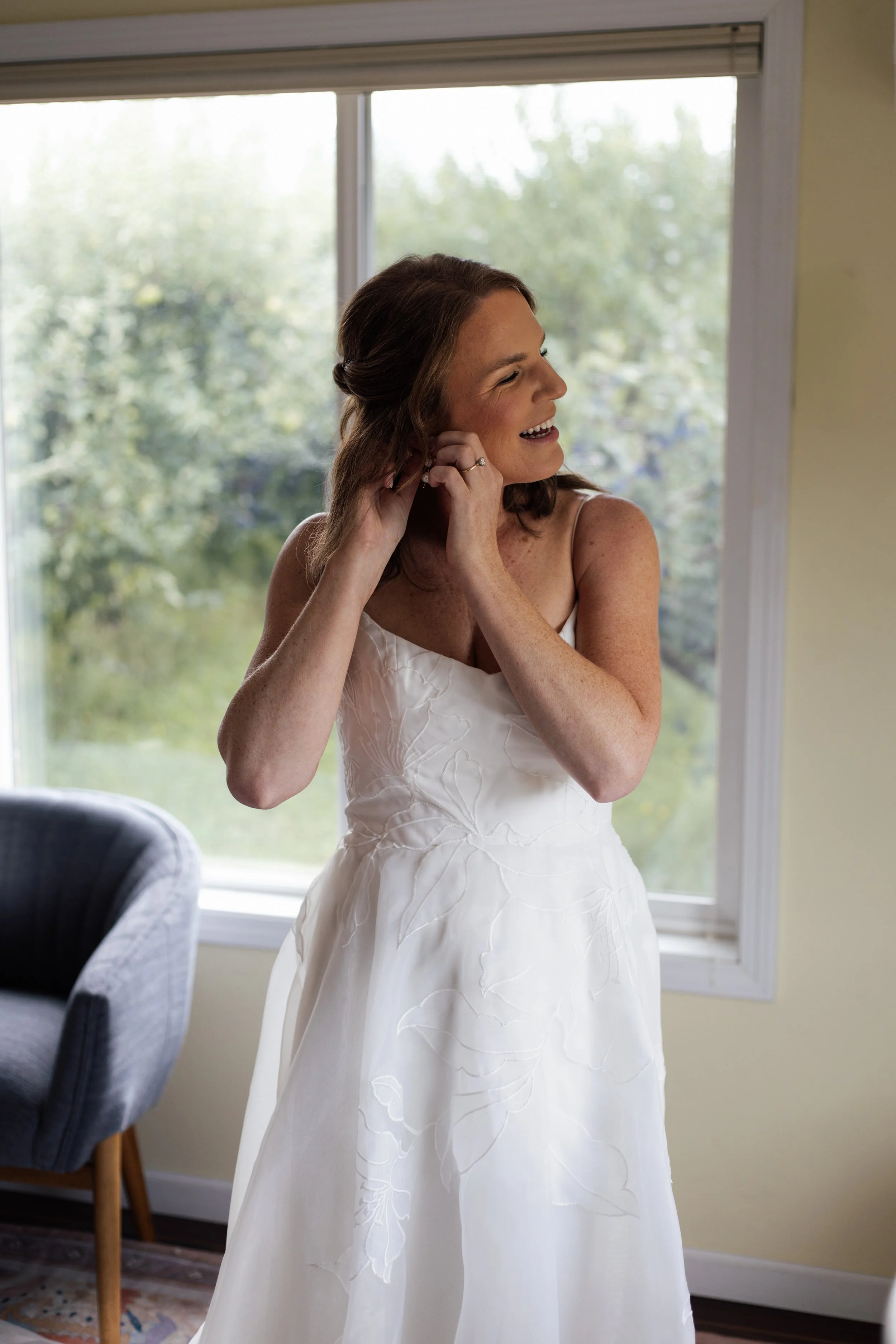 A woman in a white dress smiling while adjusting her earring near a window with a view of greenery outside.