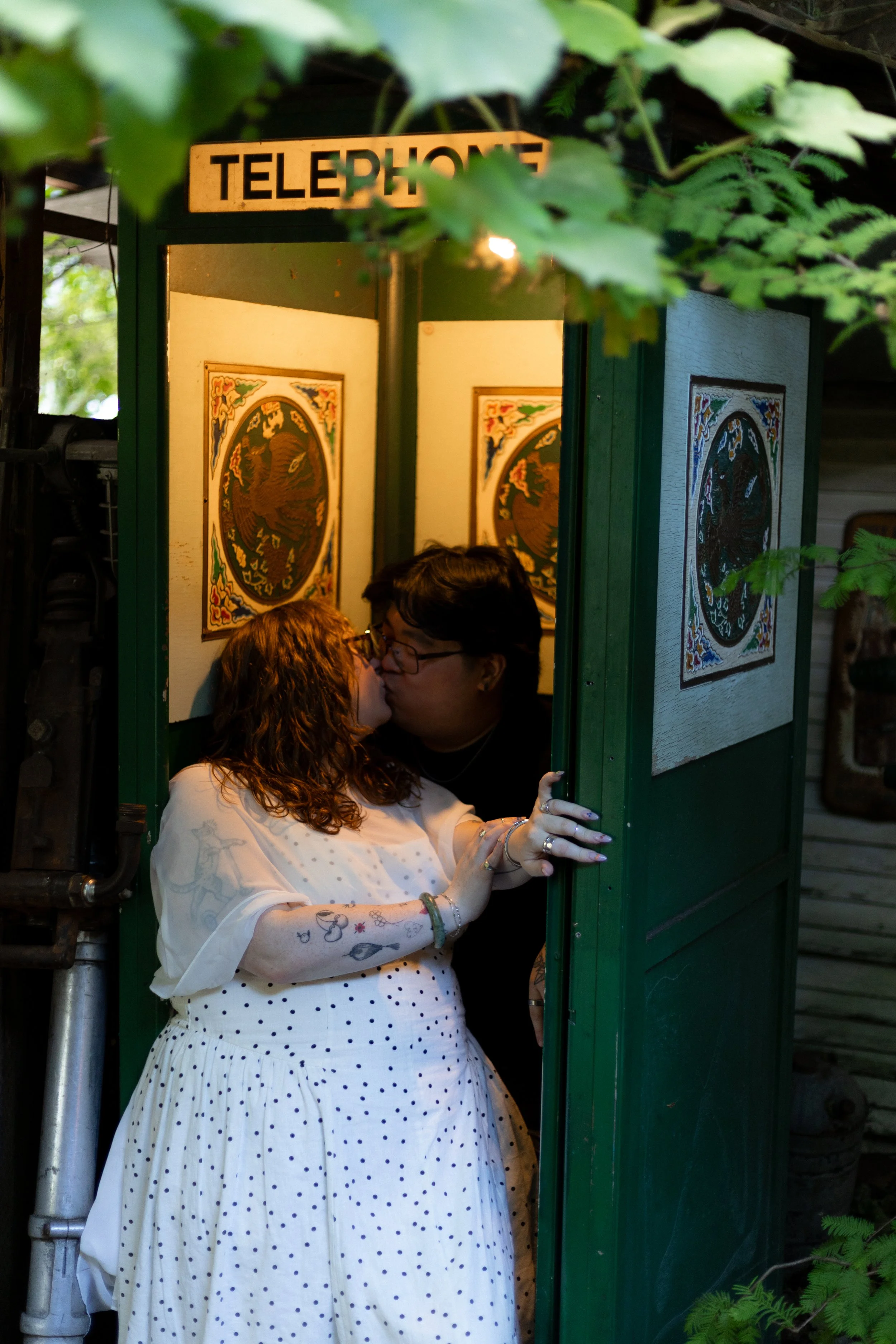 A couple kissing inside a green phone booth with vintage artwork on the walls and surrounded by greenery.