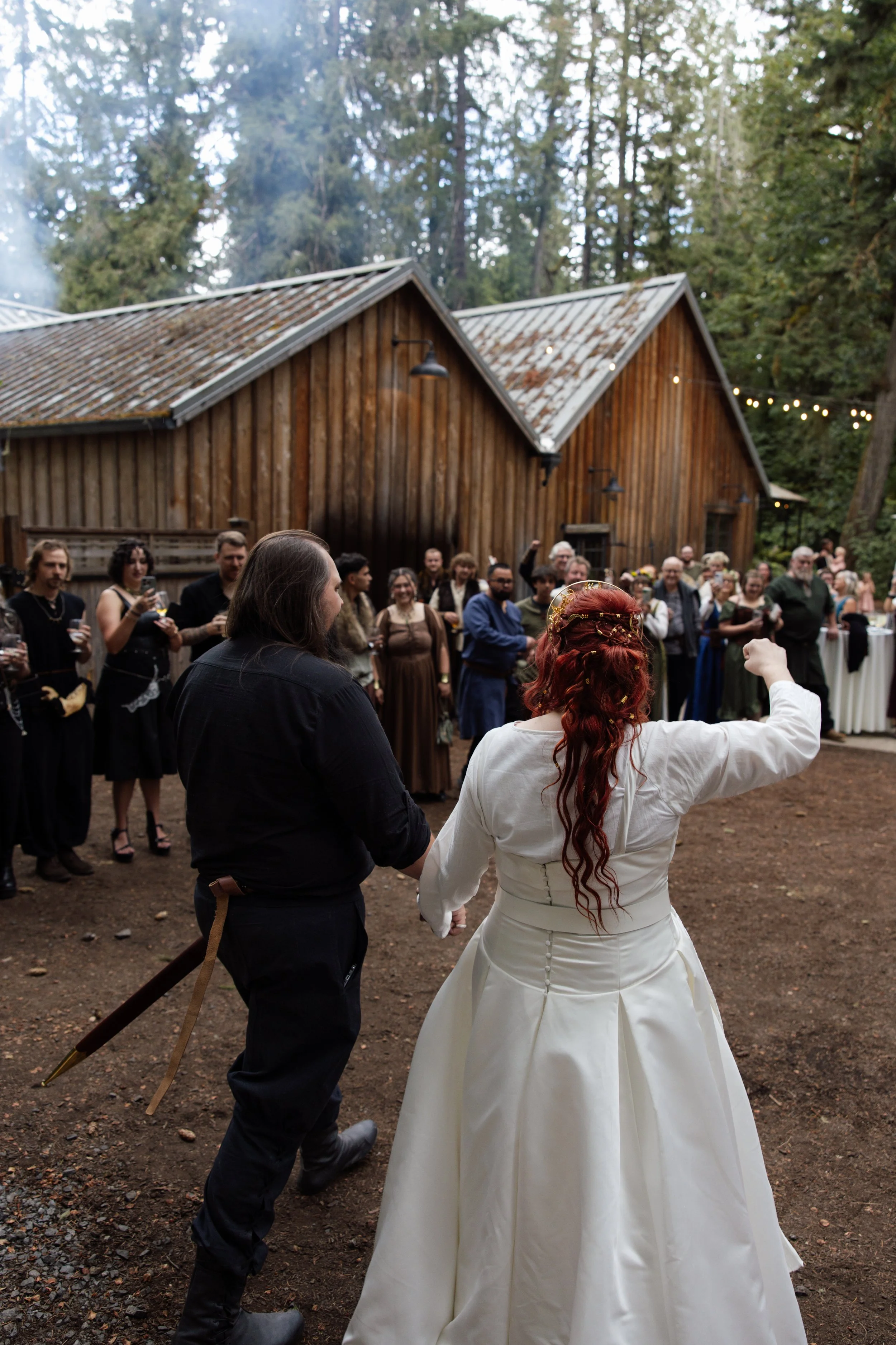 A bride and groom hold hands in a forested outdoor wedding ceremony surrounded by guests. The bride has long red hair and is dressed in a white gown, while the groom has long hair and a beard, dressed in black.