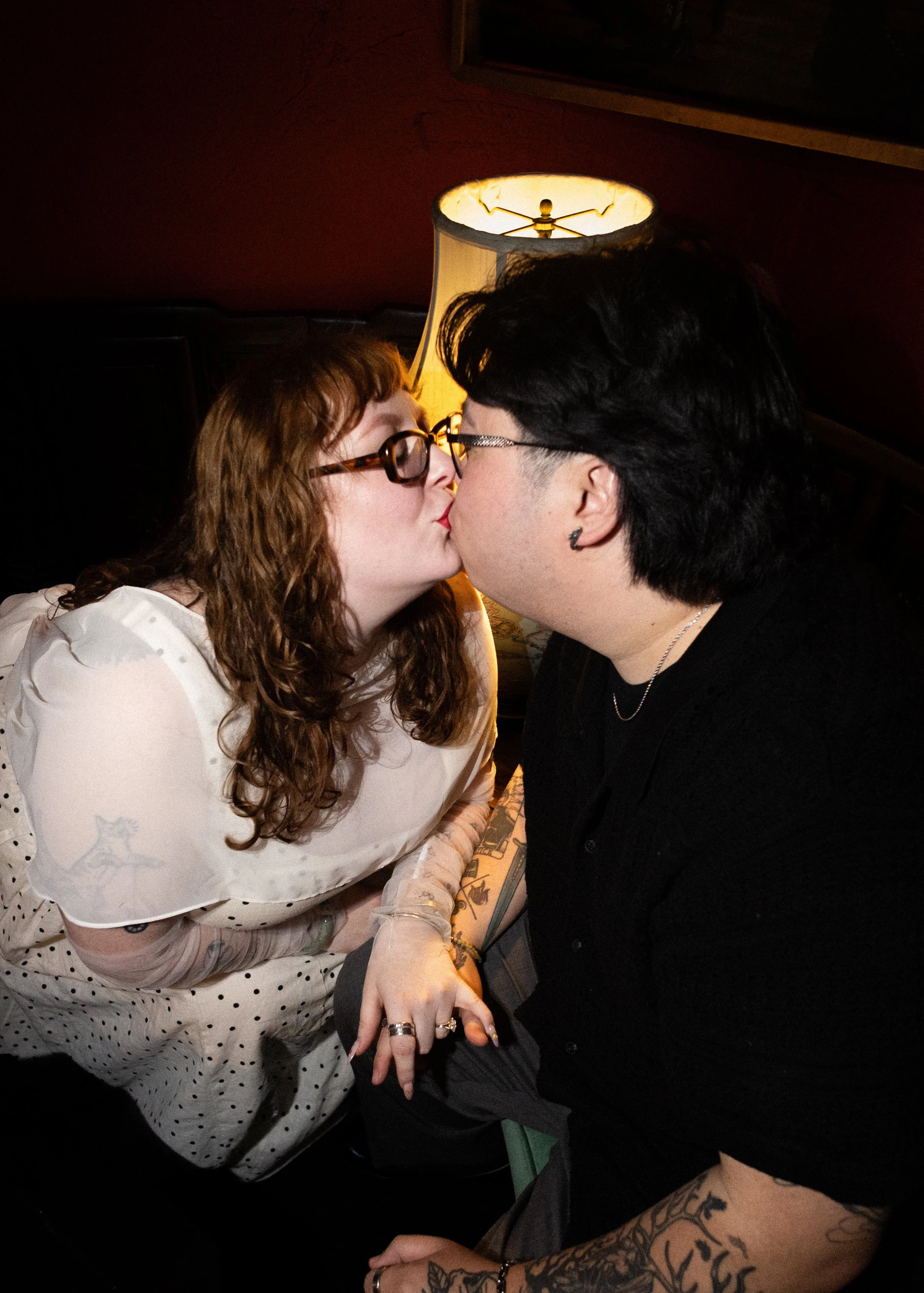 Two women are kissing. The woman on the left has curly red hair, glasses, and is wearing a white polka dot dress. The woman on the right has short dark hair, glasses, and visible tattoos on her arm. They are holding hands and seated close to each oth