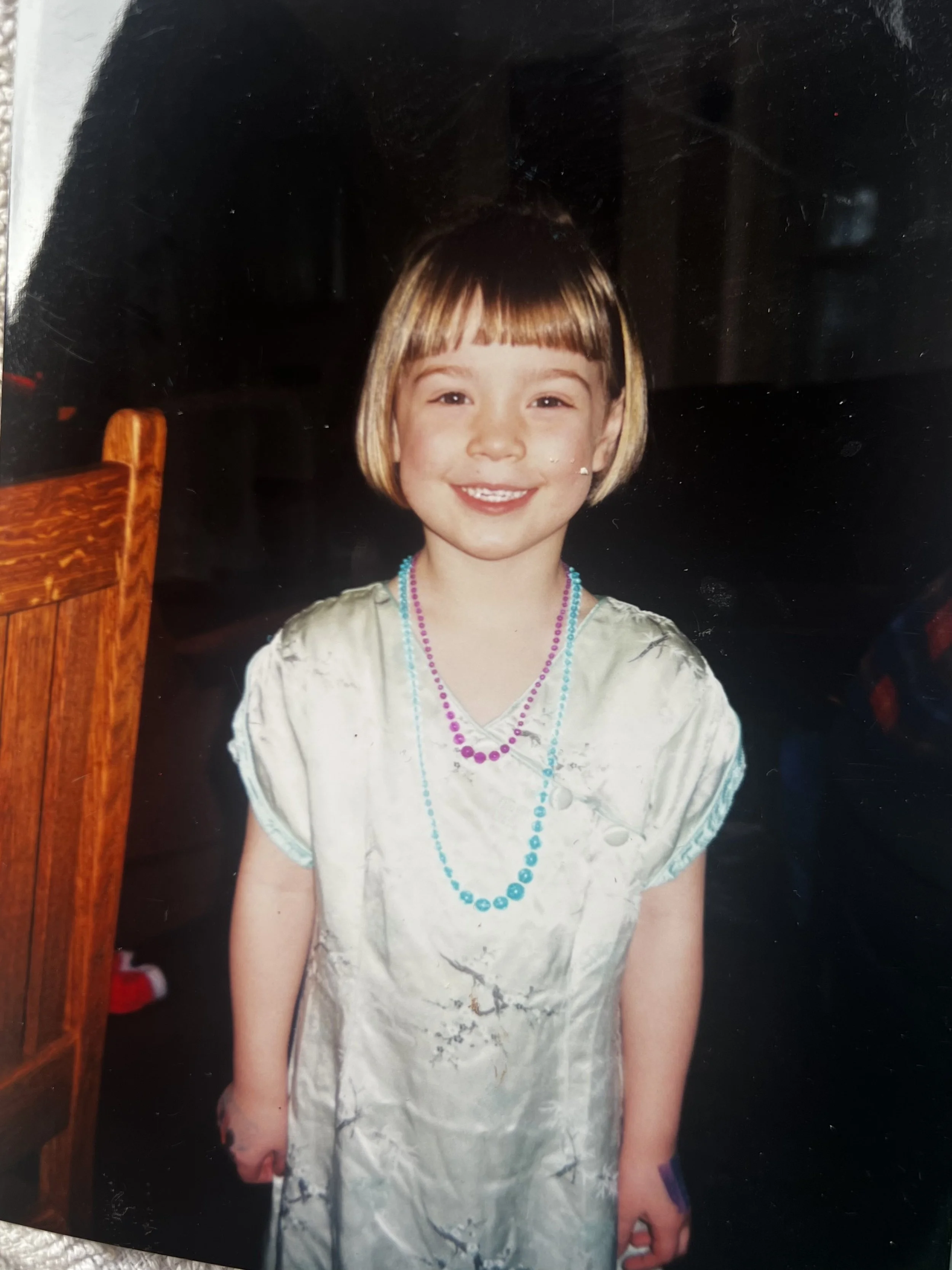 A young girl with a bob haircut and straight bangs smiling, wearing a shiny dress and multiple colorful bead necklaces, standing indoors with part of a wooden chair and a dark background visible.