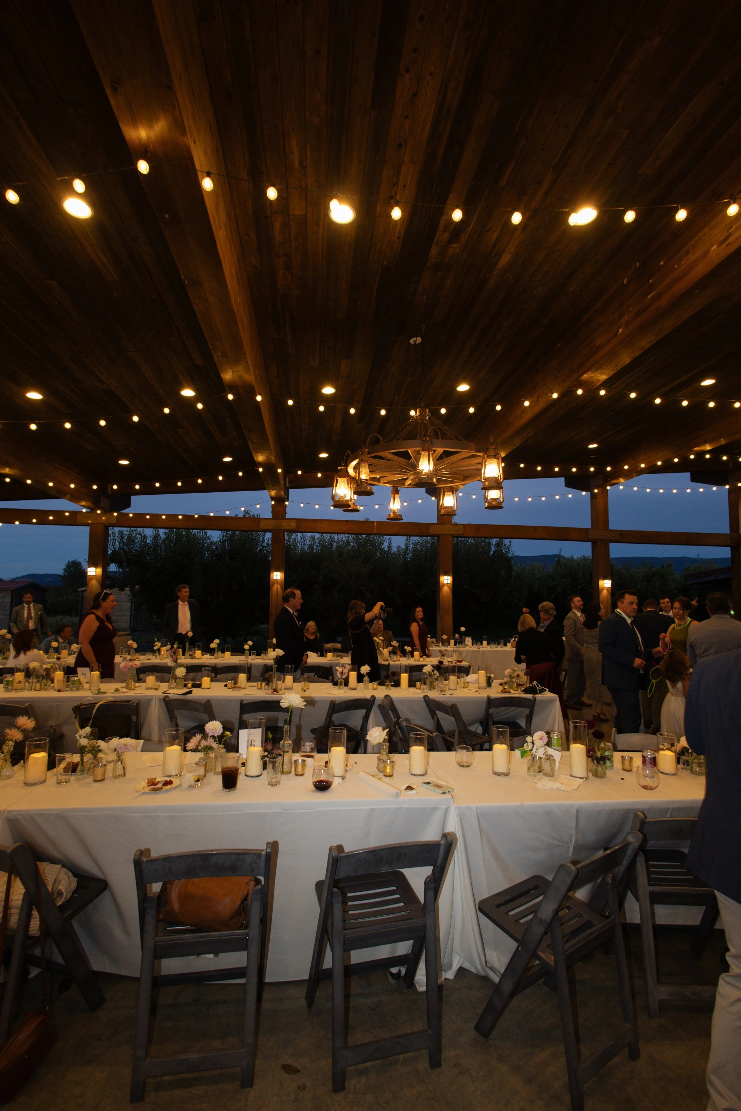 Outdoor wedding reception with decorated tables, string lights, and a covered patio at dusk.