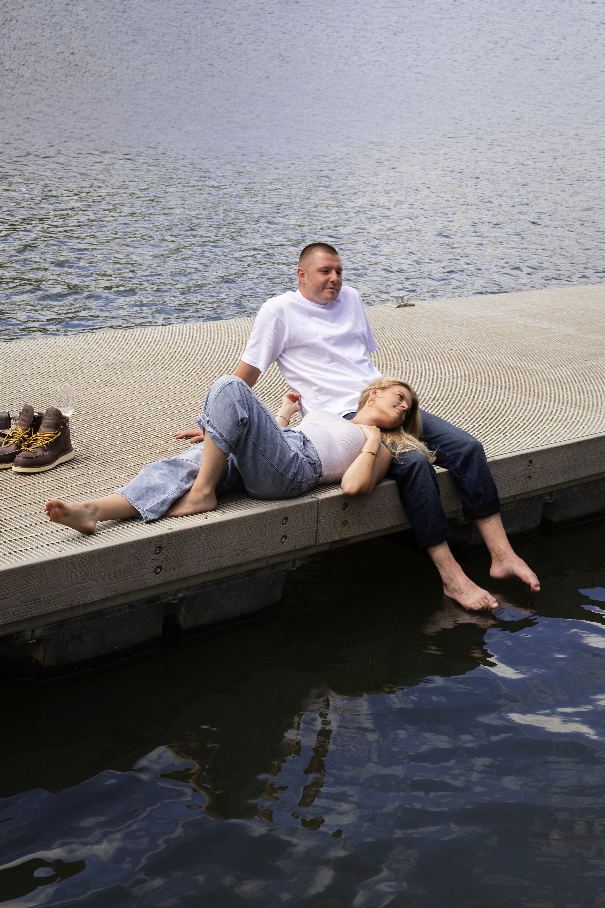 A man and a woman relaxing on a dock by a body of water, with the woman lying on the man's lap and their feet dipping in the water, shoes and a wine glass nearby.