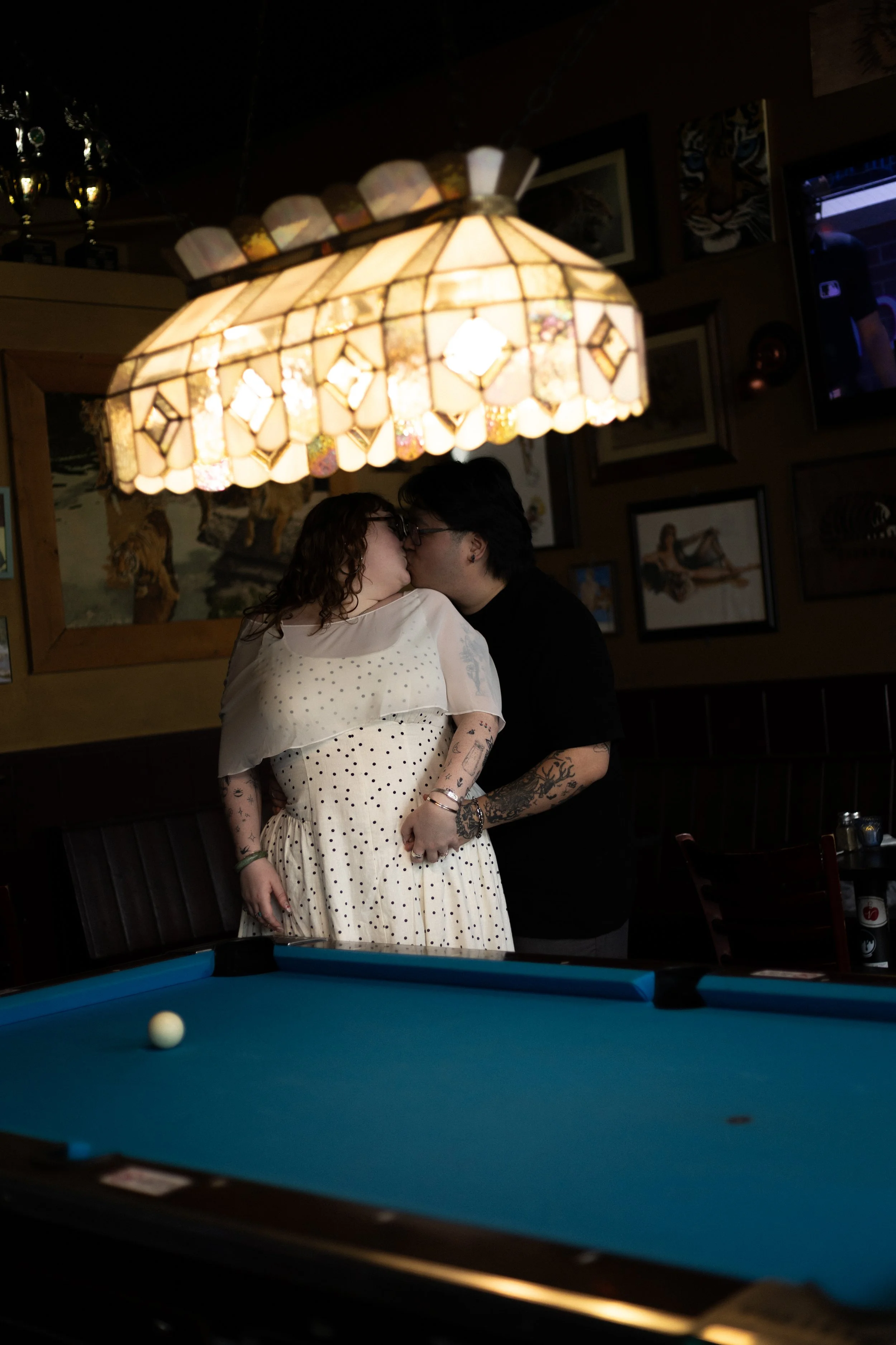 A couple sharing a kiss under a stained glass lamp in a cozy, art-filled bar or restaurant with pool table in the foreground.
