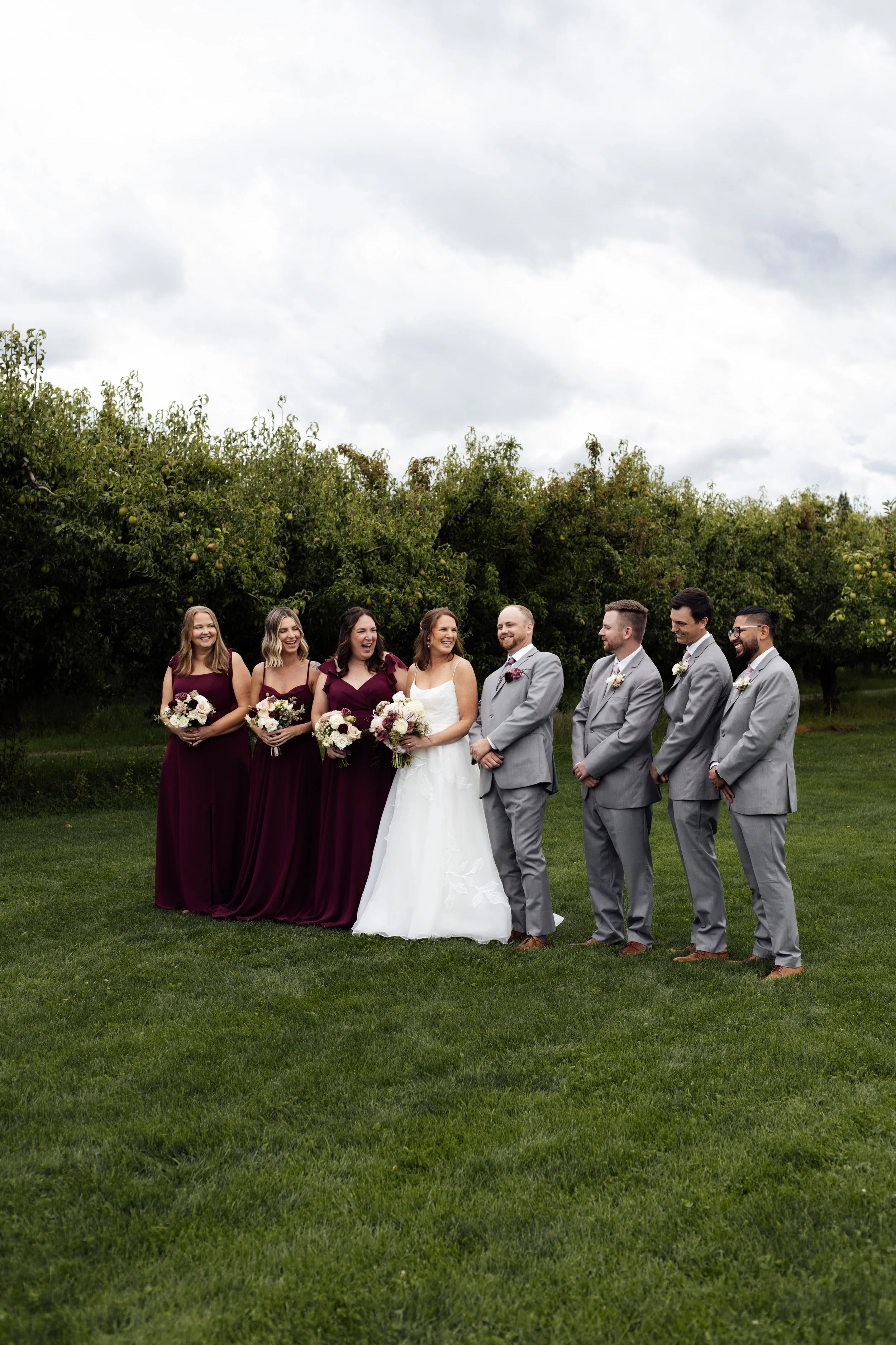 A wedding party of eight people standing outdoors on a green lawn with a backdrop of green trees and a cloudy sky. The bride is in the center wearing a white dress, holding a bouquet, and smiling. Bridesmaids on her left are in matching dark purple d