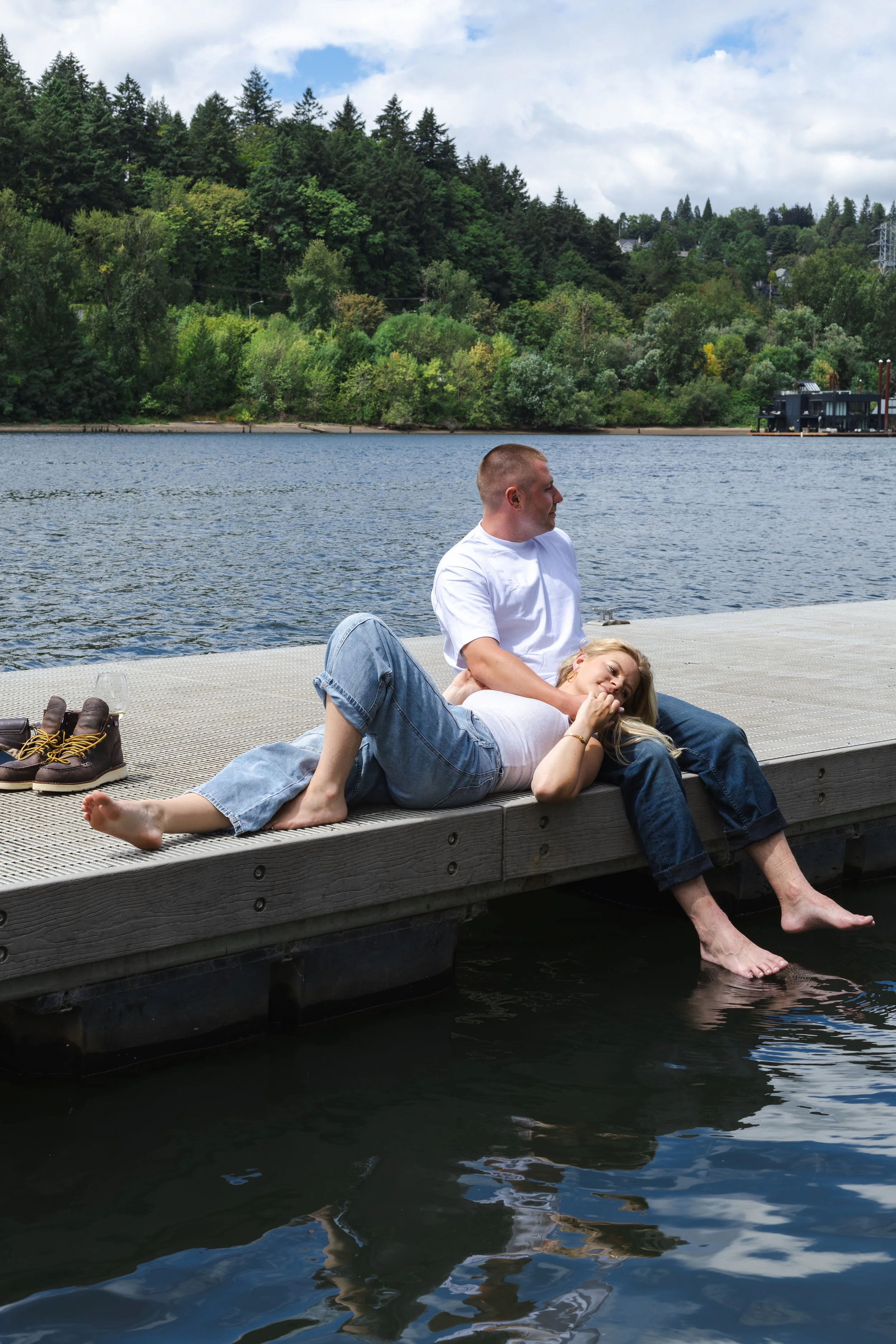 A young man and woman are relaxing on a dock by a lake, with the woman lying with her head on the man's lap, both smiling. The background features a dense green forest and a cloudy sky.