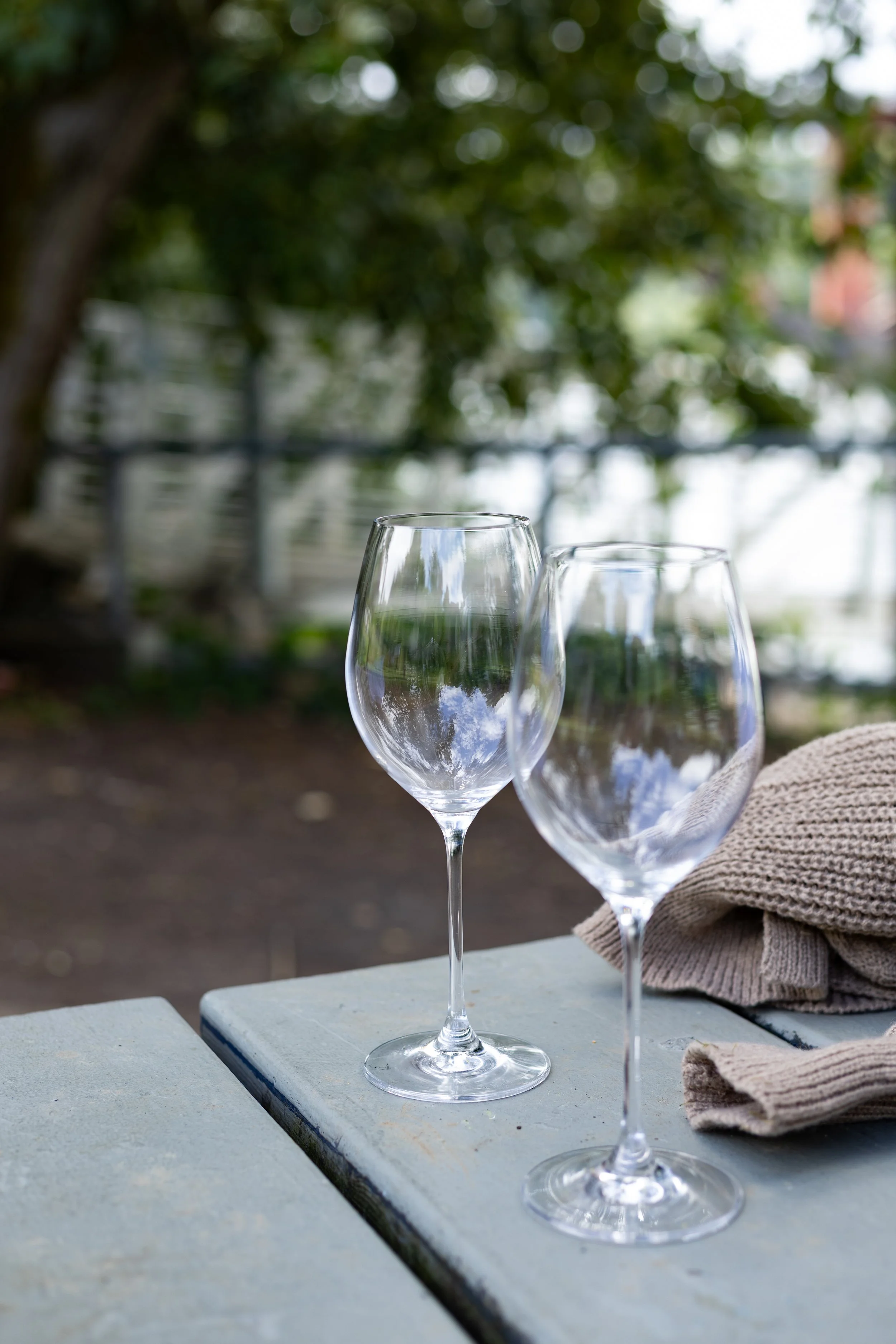 Two empty wine glasses on a white wooden table outdoors with a blurred green leafy background, a beige cloth napkin to the side.