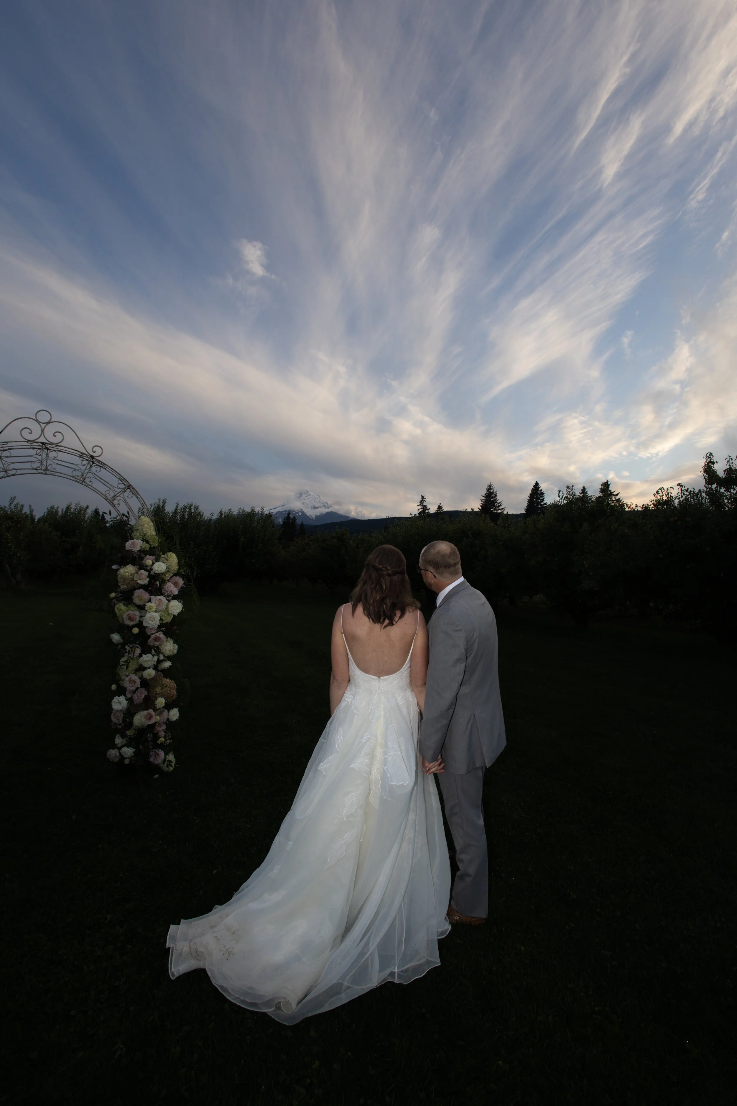 A bride and groom stand hand in hand outdoors during their wedding ceremony at sunset, with a scenic mountain and cloudy sky in the background.