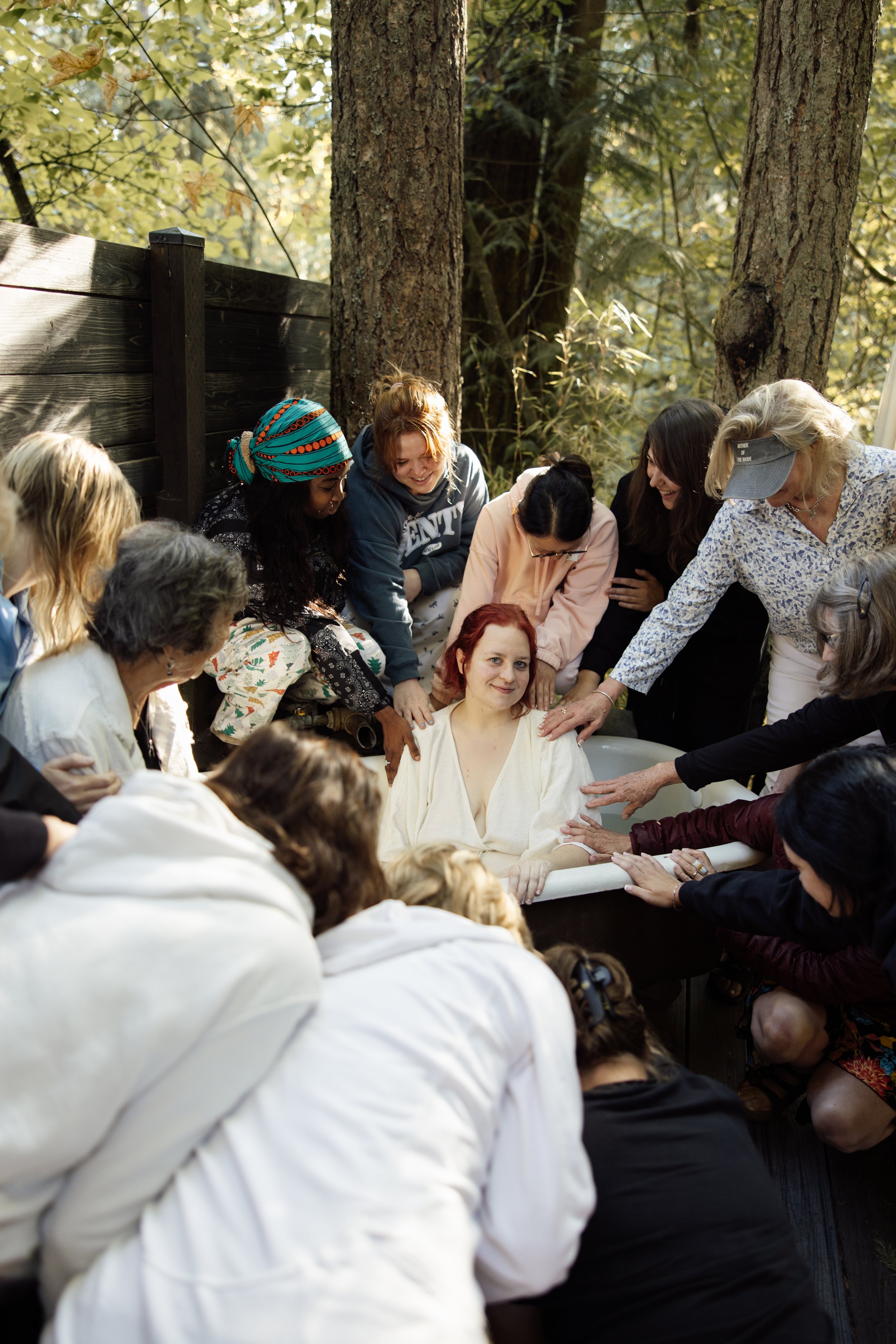 A group of women gathered outdoors around a woman seated in a bathtub, with trees and sunlight in the background. They appear to be participating in a celebration or ritual.