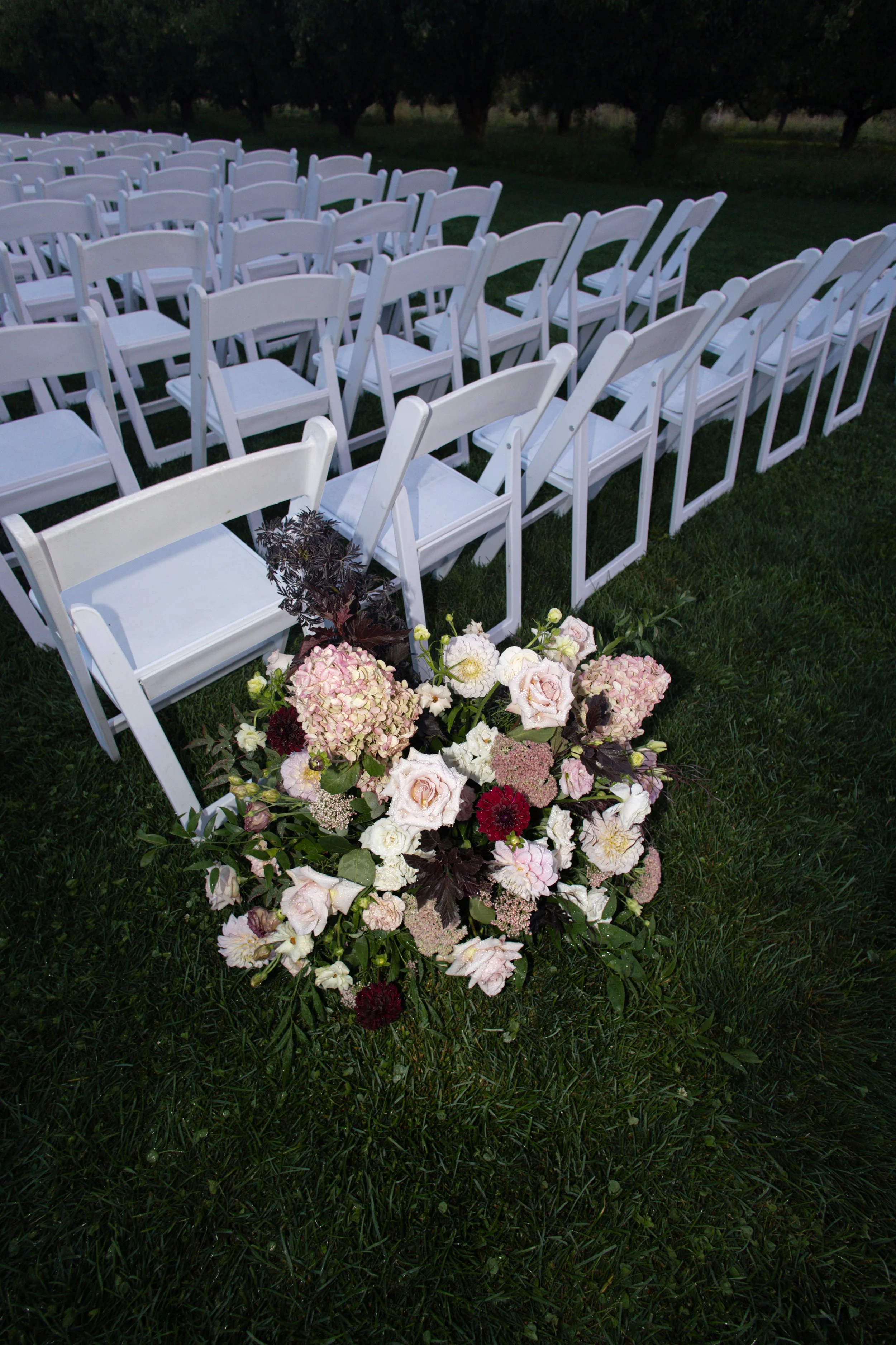 White folding chairs arranged outdoors on grass with a floral arrangement in the foreground, set for an event.