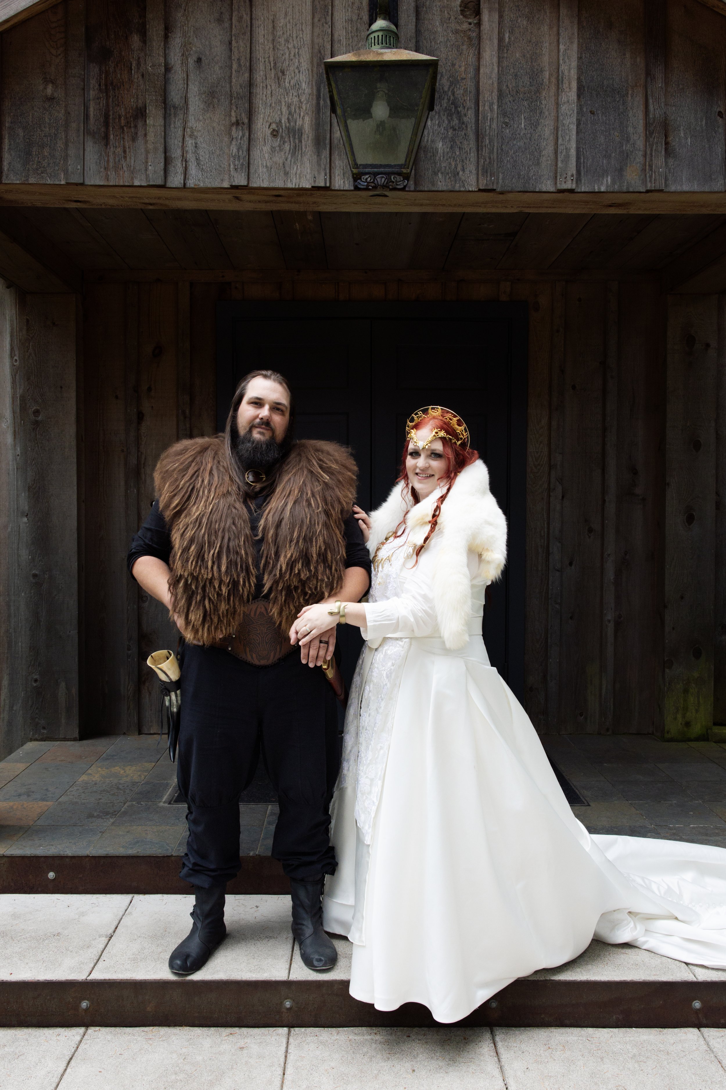 A man and a woman in medieval-inspired costumes standing together outside in front of a wooden structure with a lantern above them. The man is wearing a fur vest over black clothing, and the woman is dressed in a white gown with a fur cape and a decorative gold crown.