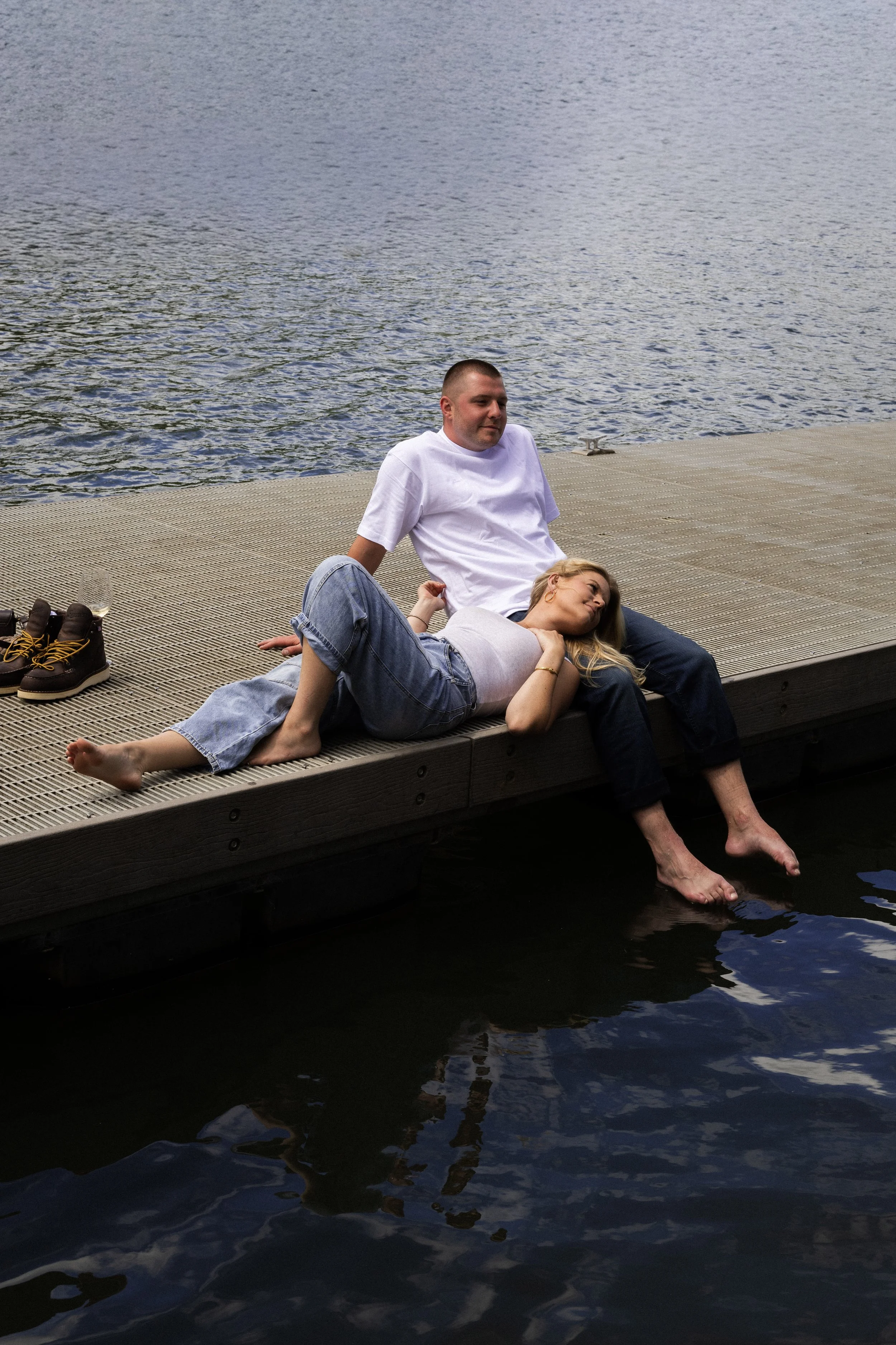 Two people relaxing on a wooden dock by a lake, with one person lying down and the other sitting up, feet dipped in the water, shoes and a wine glass nearby, overcast sky.