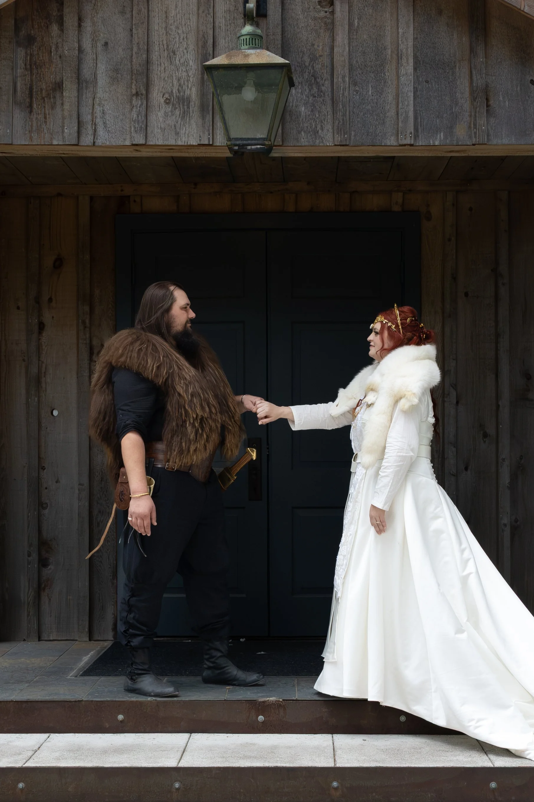 A man and woman dressed in medieval or fantasy-style costumes, holding hands and facing each other in front of a dark door with wooden paneling. The man has long hair and a beard, wearing black clothing with a fur vest, and the woman has red hair, we