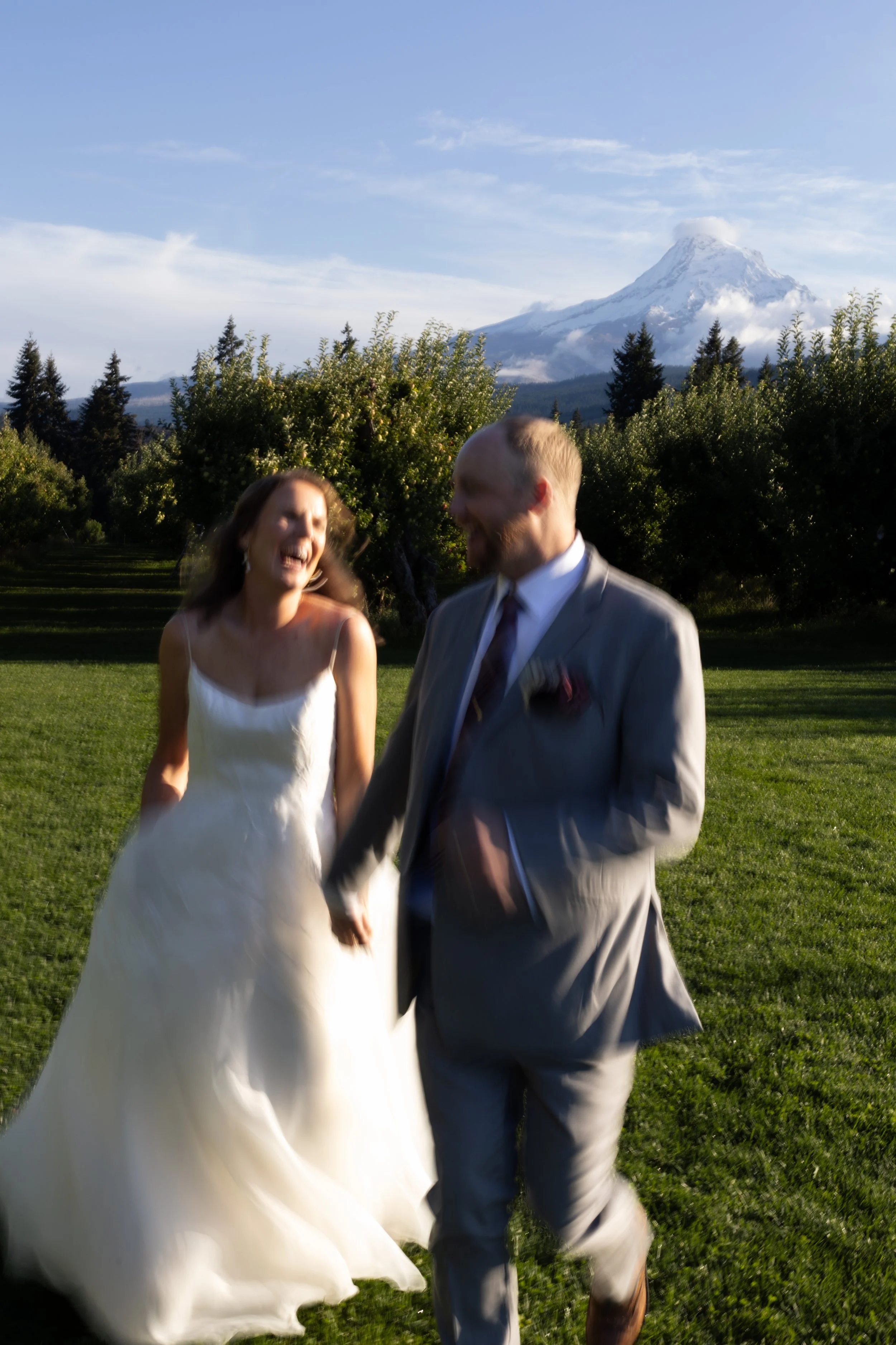 A couple dancing outdoors in a grassy area with trees and Mount Hood in the background. The woman is wearing a white dress and the man is dressed in a gray suit.