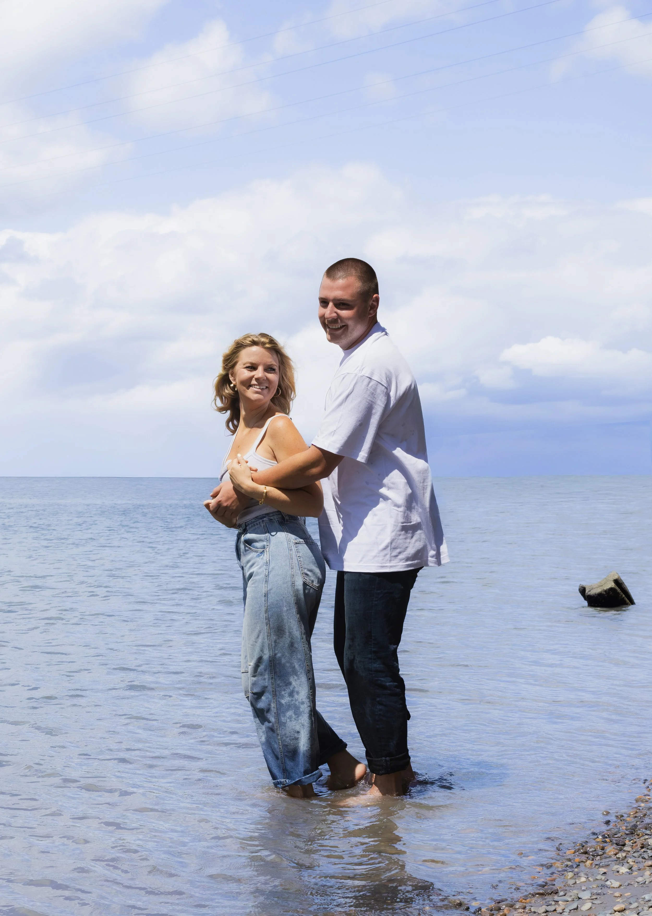 A couple standing in shallow water at the beach, smiling, with a cloudy sky above and some rocks on the shoreline.