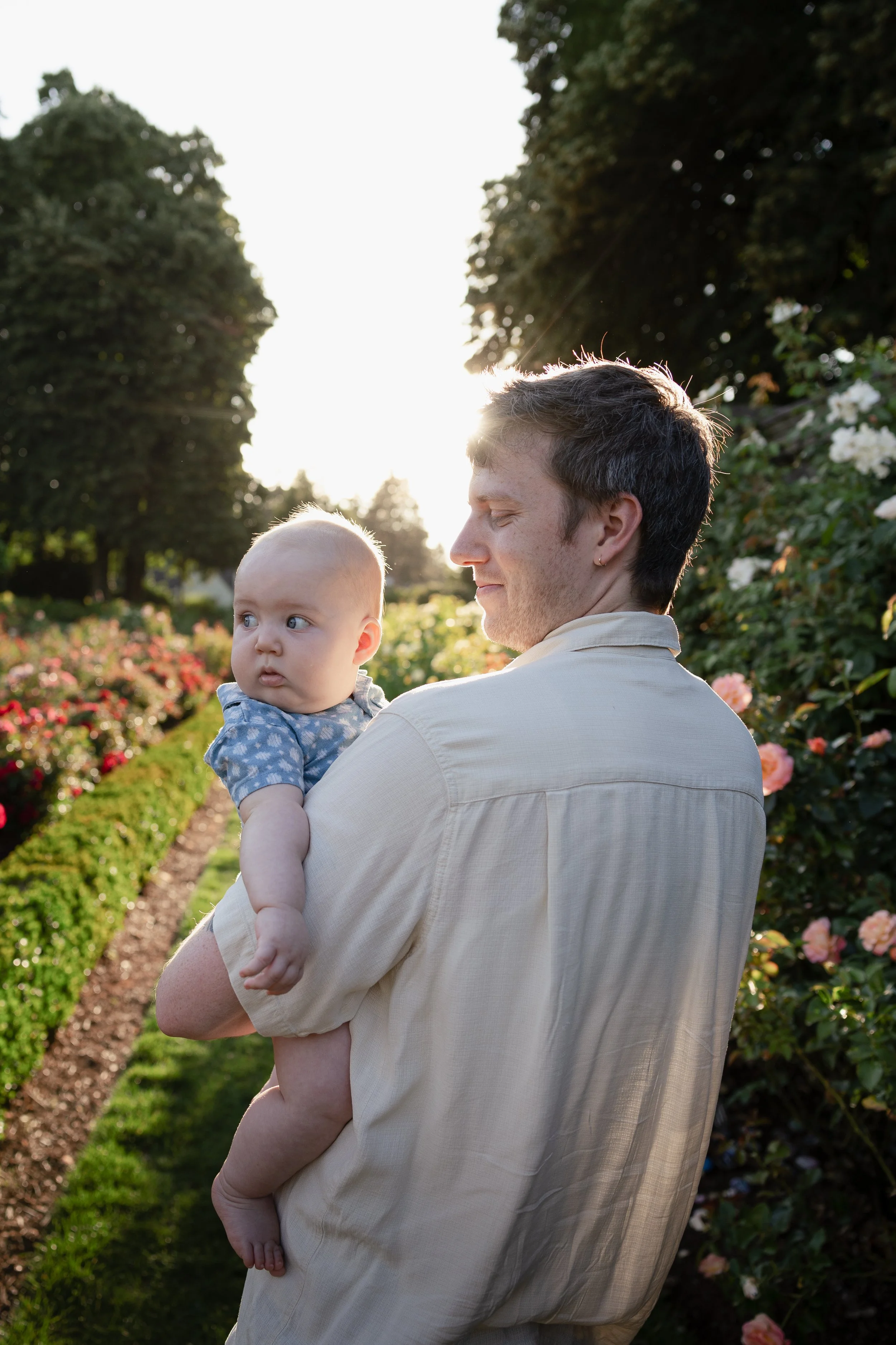 A man with dark hair and light skin holding a baby with light skin and light-colored hair in a garden with pink and white flowers, green bushes, and trees, with the sun setting in the background.