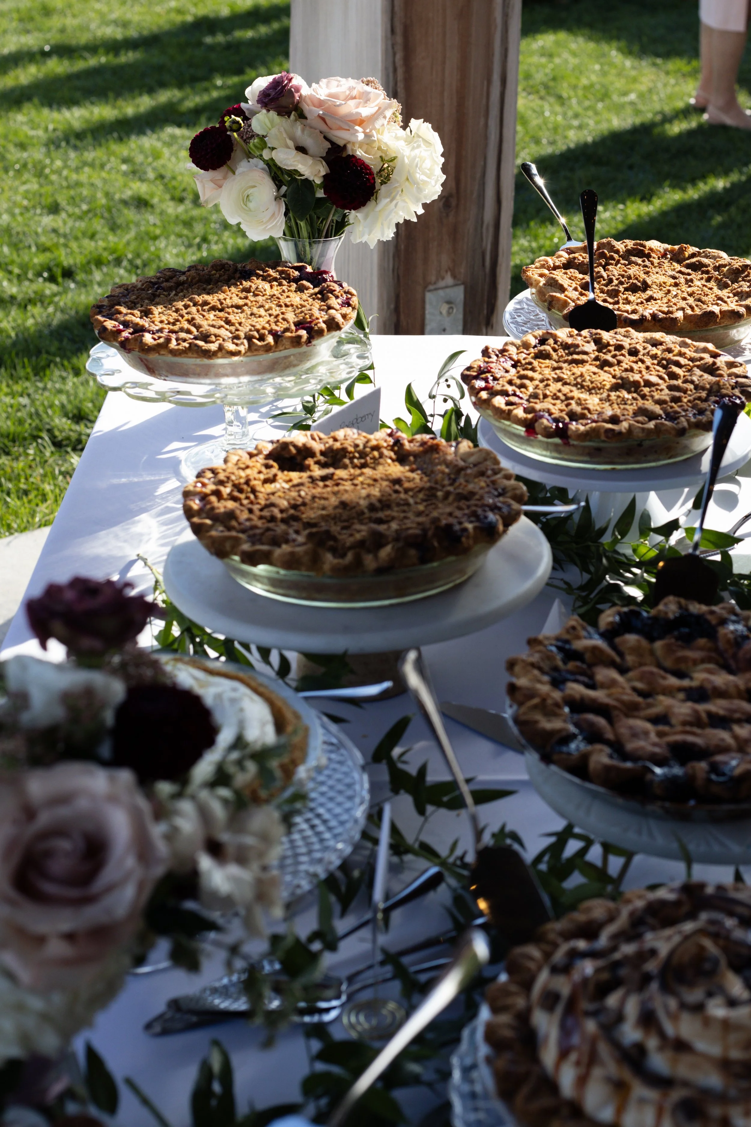 Several apple pies with crumb toppings on a white tablecloth, in glass dishes, outdoors on grass with green background and people standing nearby.