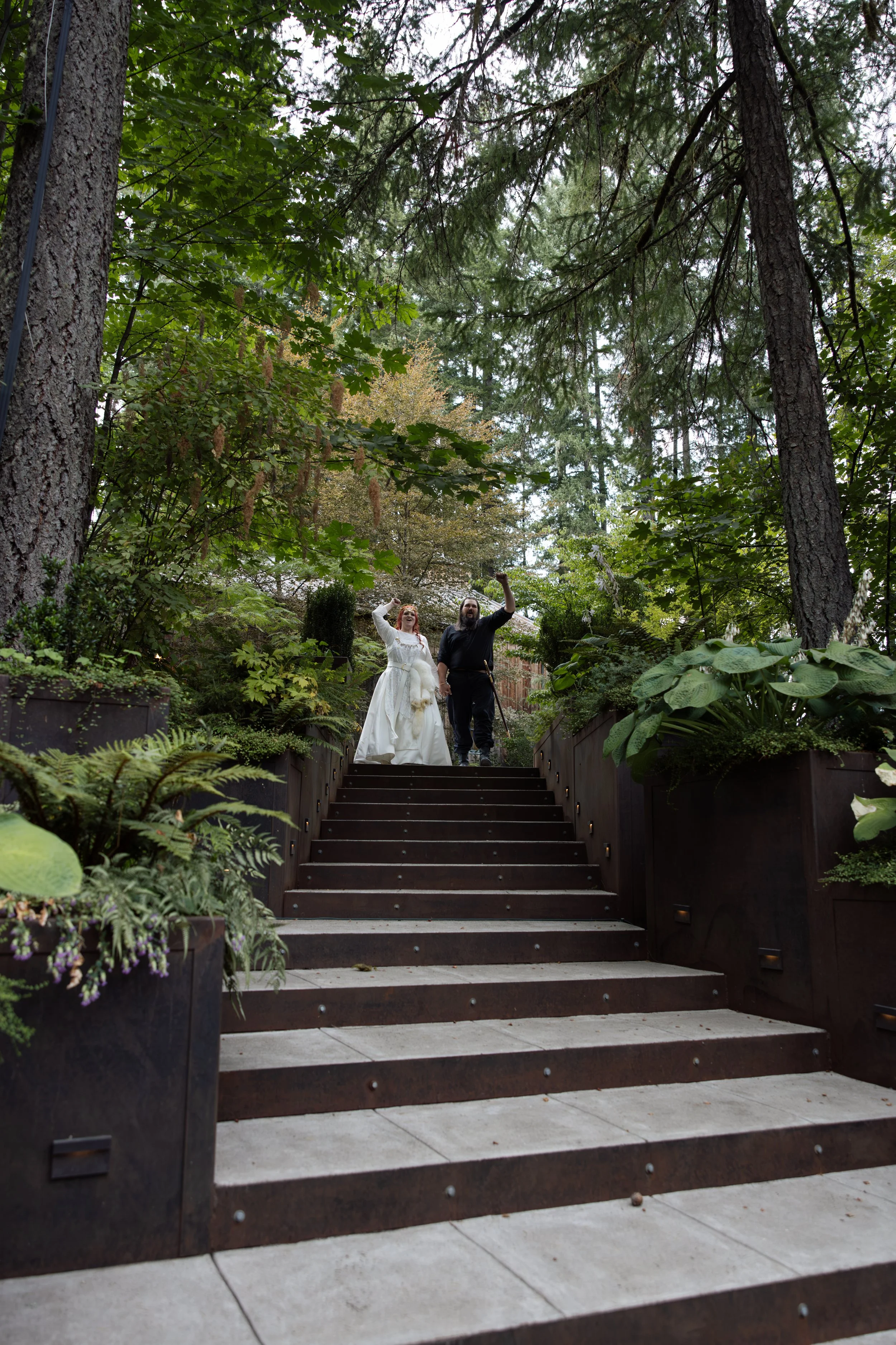 A bride and groom walking up an outdoor staircase surrounded by lush greenery in a garden setting.