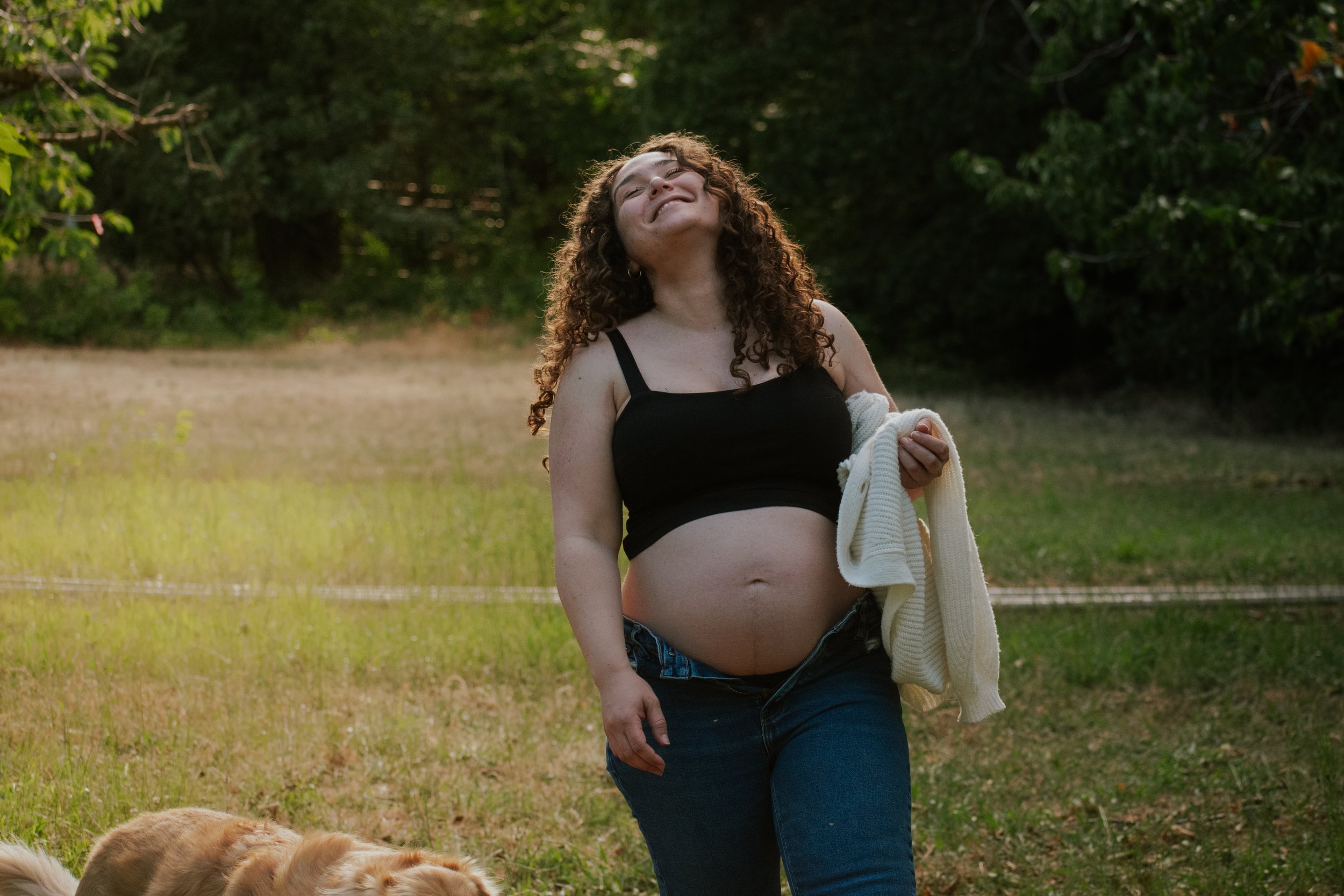A pregnant woman with curly hair smiling outdoors holding a white sweater, with a golden retriever lying on the grass nearby and green trees in the background.
