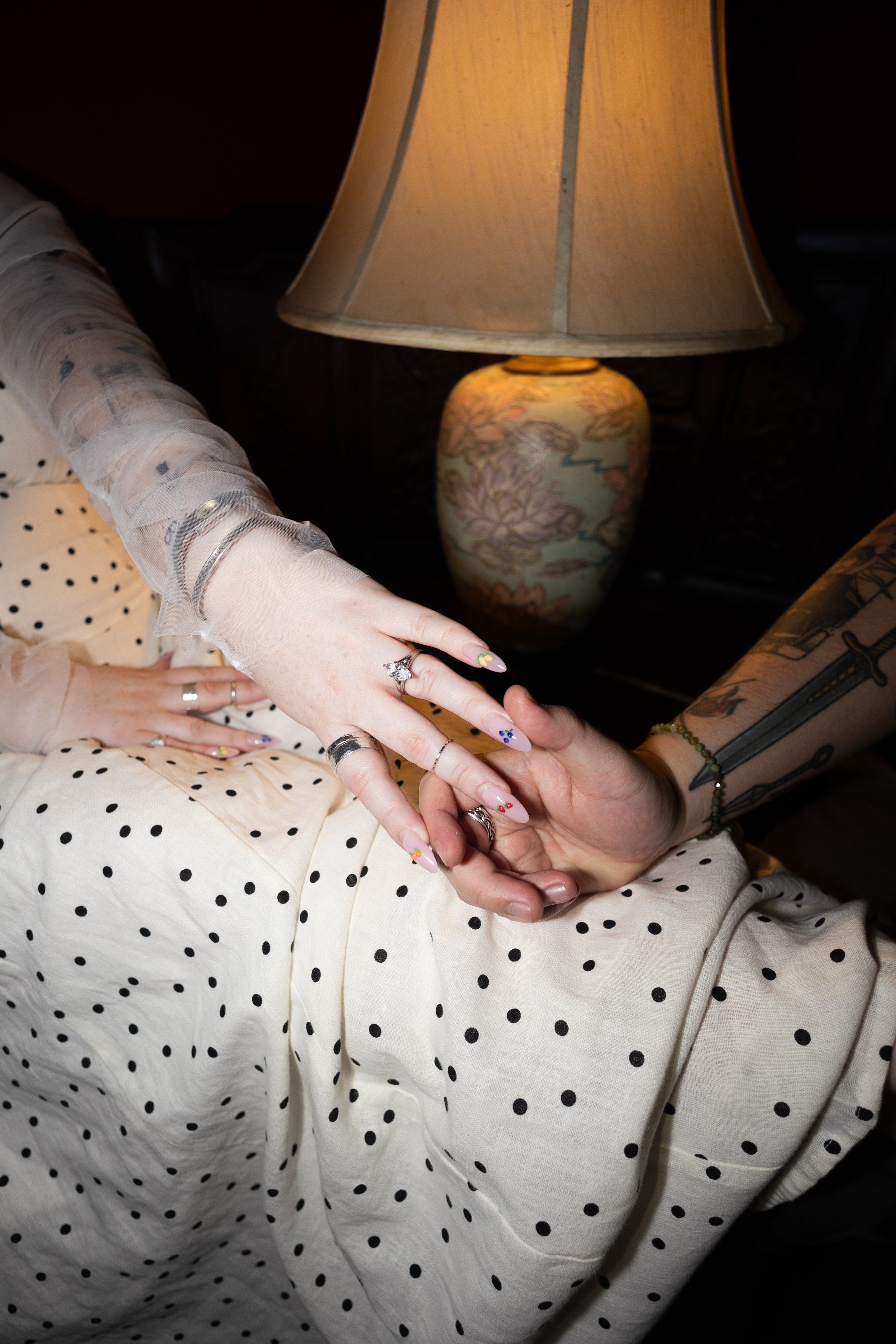 Close-up of two people holding hands, one person with decorated nails and multiple rings; the other person with a tattooed arm and bracelet, wearing a polka-dot shirt. Background includes a lamp on a wooden table.