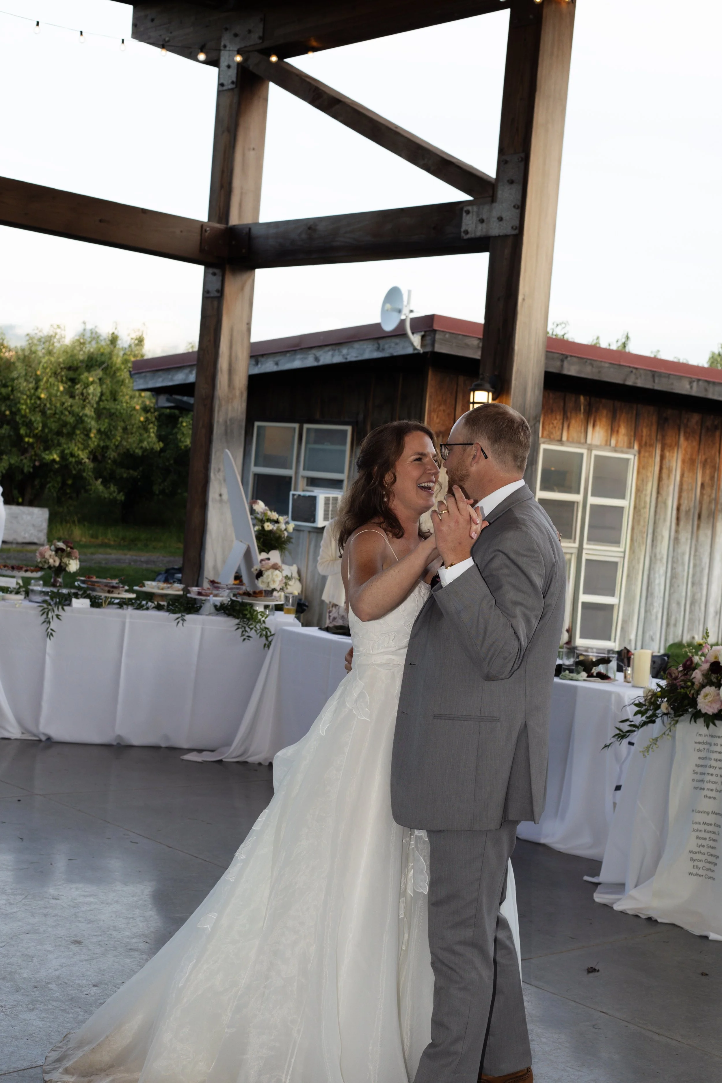 A bride and groom dancing at their wedding reception outdoors during the evening. The bride is wearing a white wedding gown, and the groom is in a light gray suit. They are smiling and looking at each other, holding hands. There is a decorated table 