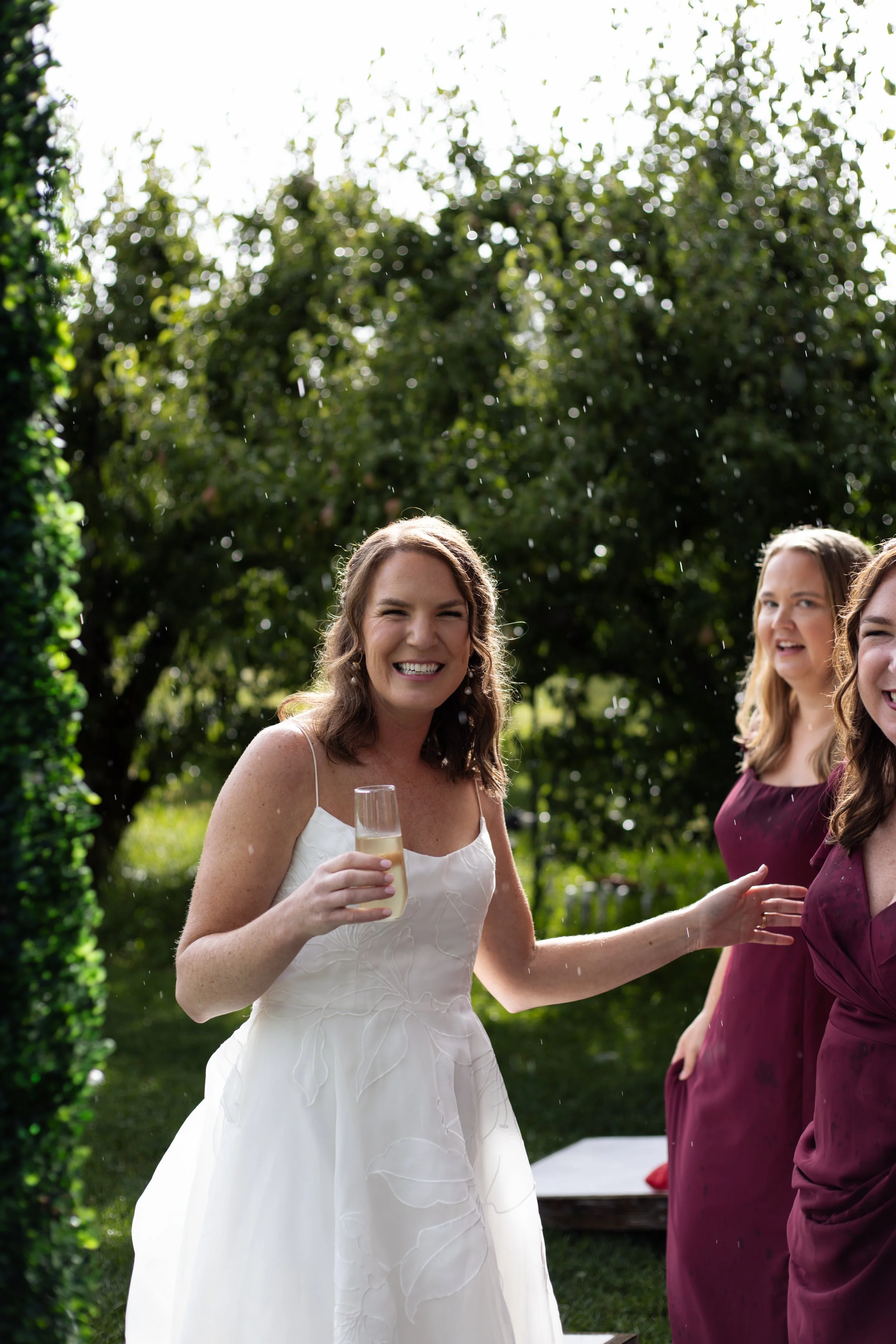 A woman in a white dress holding a champagne glass at an outdoor celebration, smiling and surrounded by other women, with greenery and trees in the background.