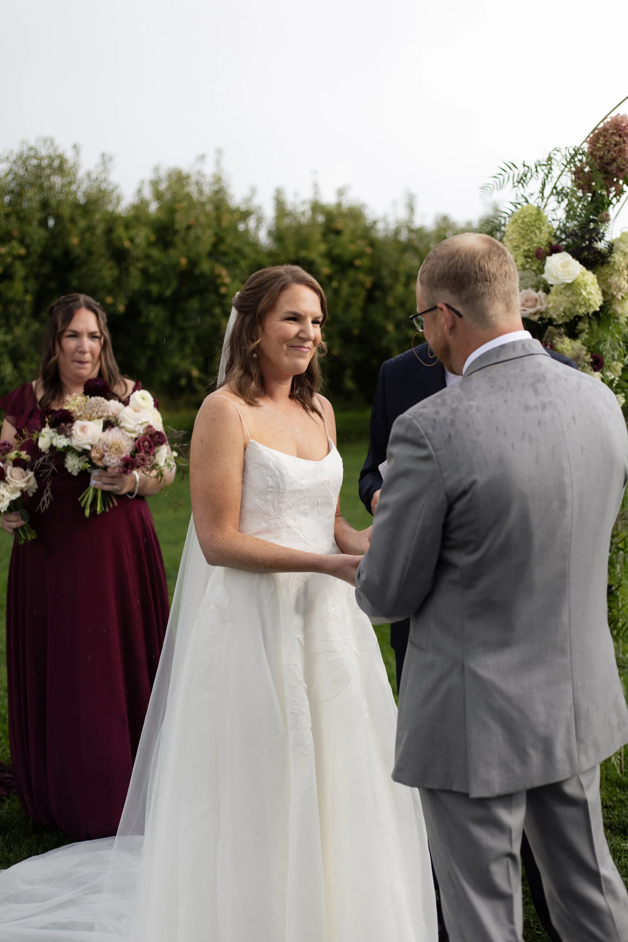 A bride with brown hair and a white dress standing outside during her wedding ceremony, smiling at her partner, with a bridesmaid holding a bouquet in the background and a floral arch behind them.