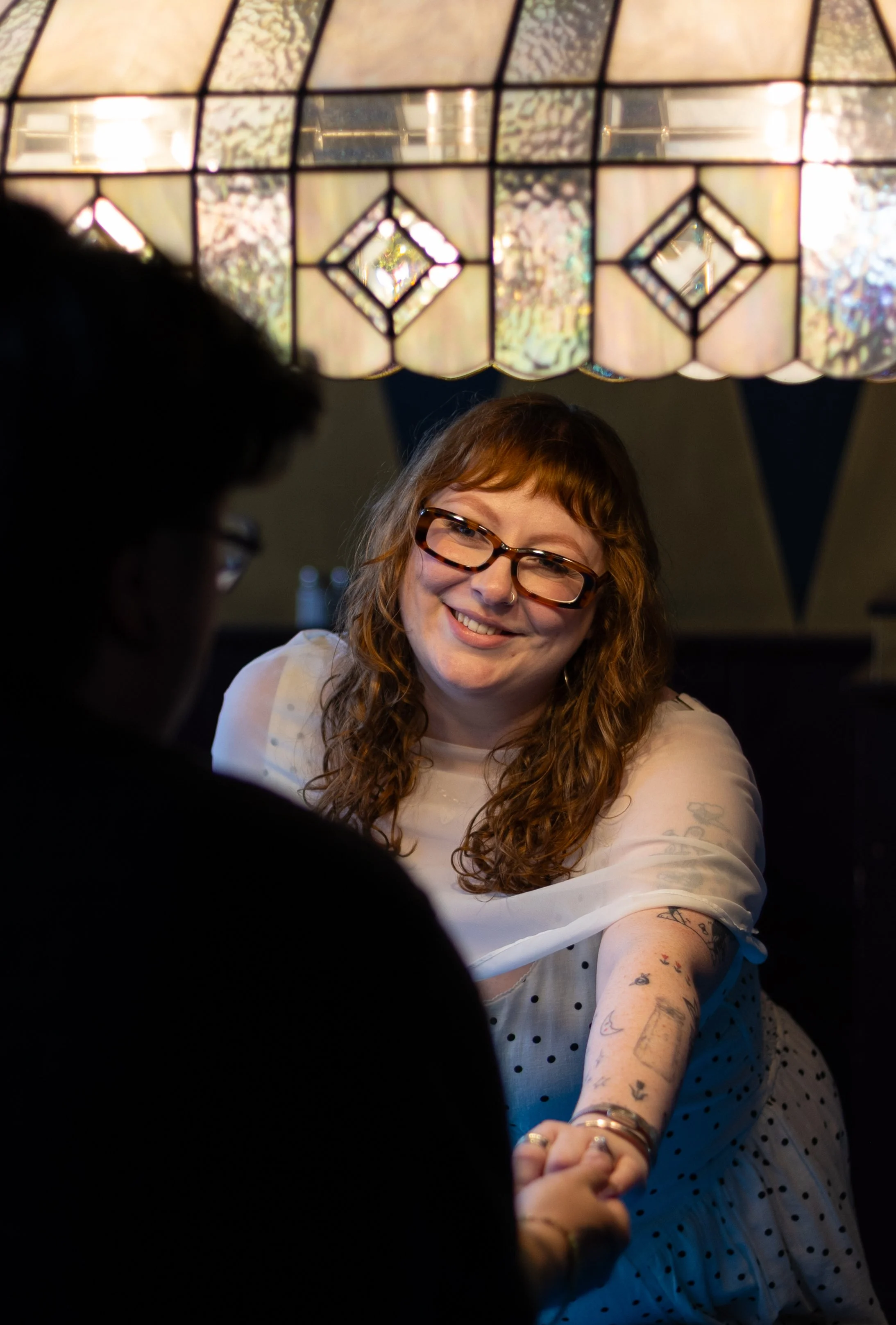 A young woman with red hair, glasses, and tattoos smiling and holding hands with someone out of the frame, under a stained glass ceiling.