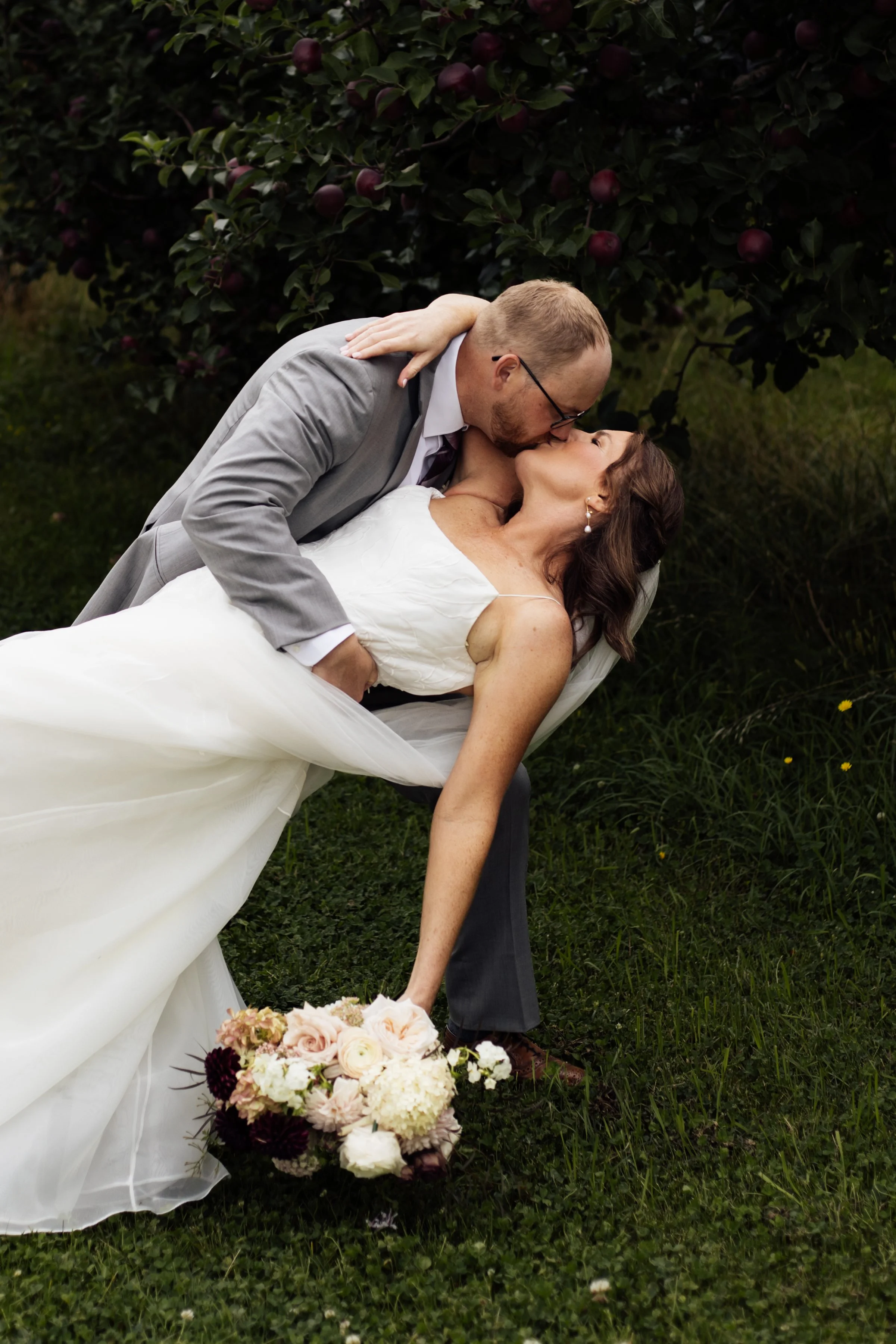 A newlywed couple sharing a kiss outdoors, with the man leaning over the woman, who is lying on the grass. The woman is holding a bouquet of flowers, and the background features a bush with red berries.