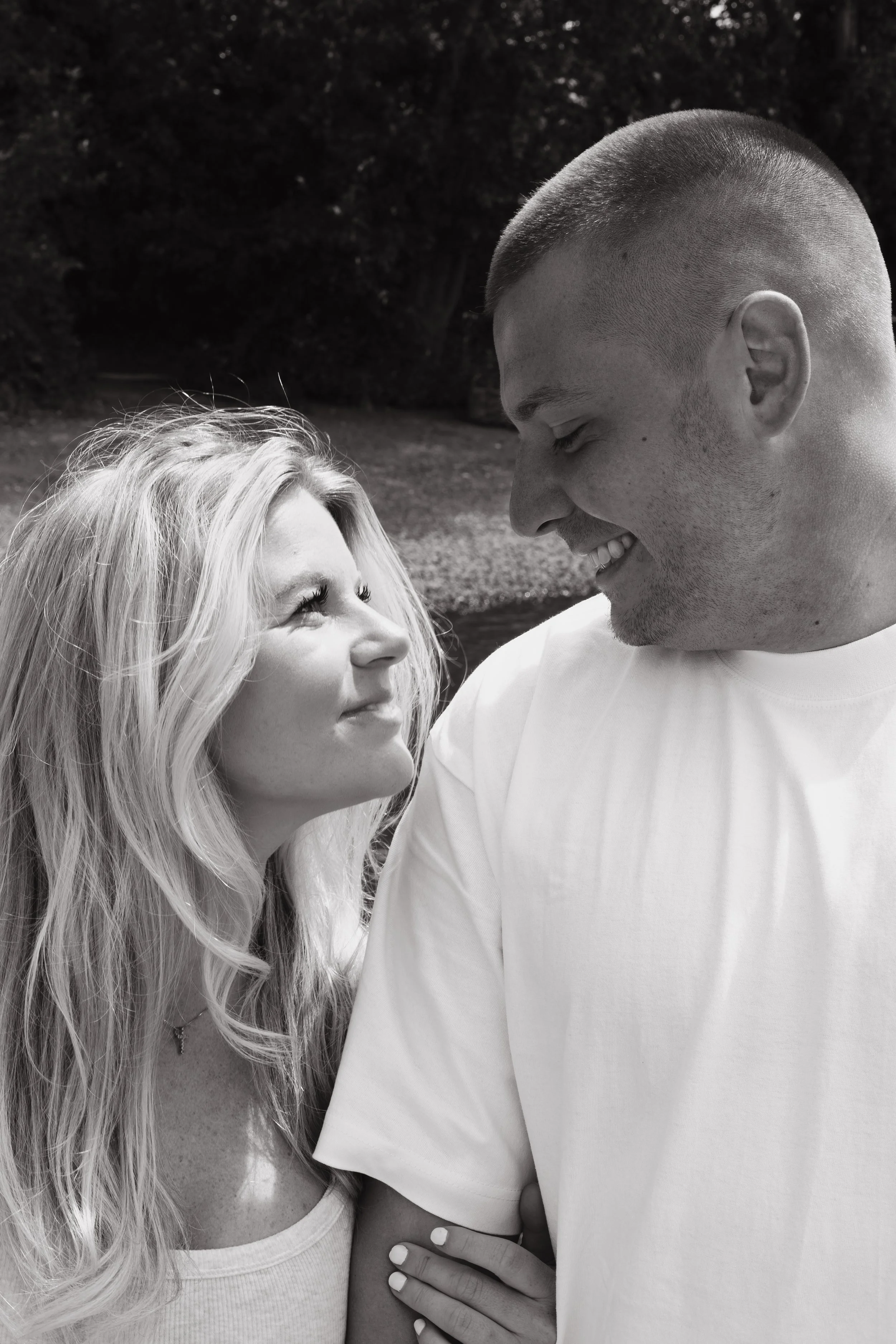 A black and white photo of a man and woman looking at each other, smiling. The woman has long, wavy blonde hair and the man has a buzzed haircut. The woman is wearing a sleeveless top and a necklace, and the man is wearing a white T-shirt. The woman 