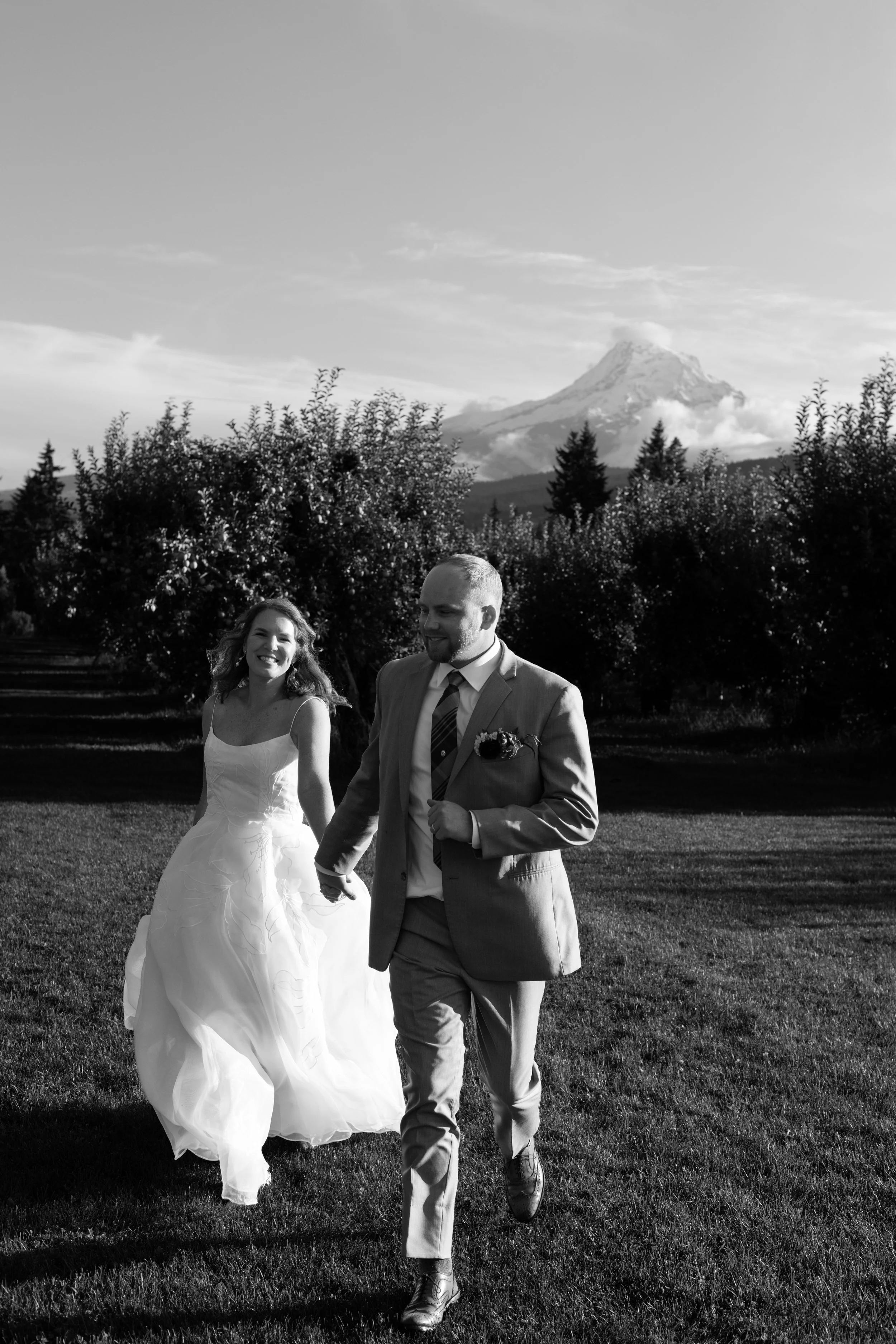 A bride and groom walking outdoors with Mount Hood in the background, smiling and holding hands on a grassy field.